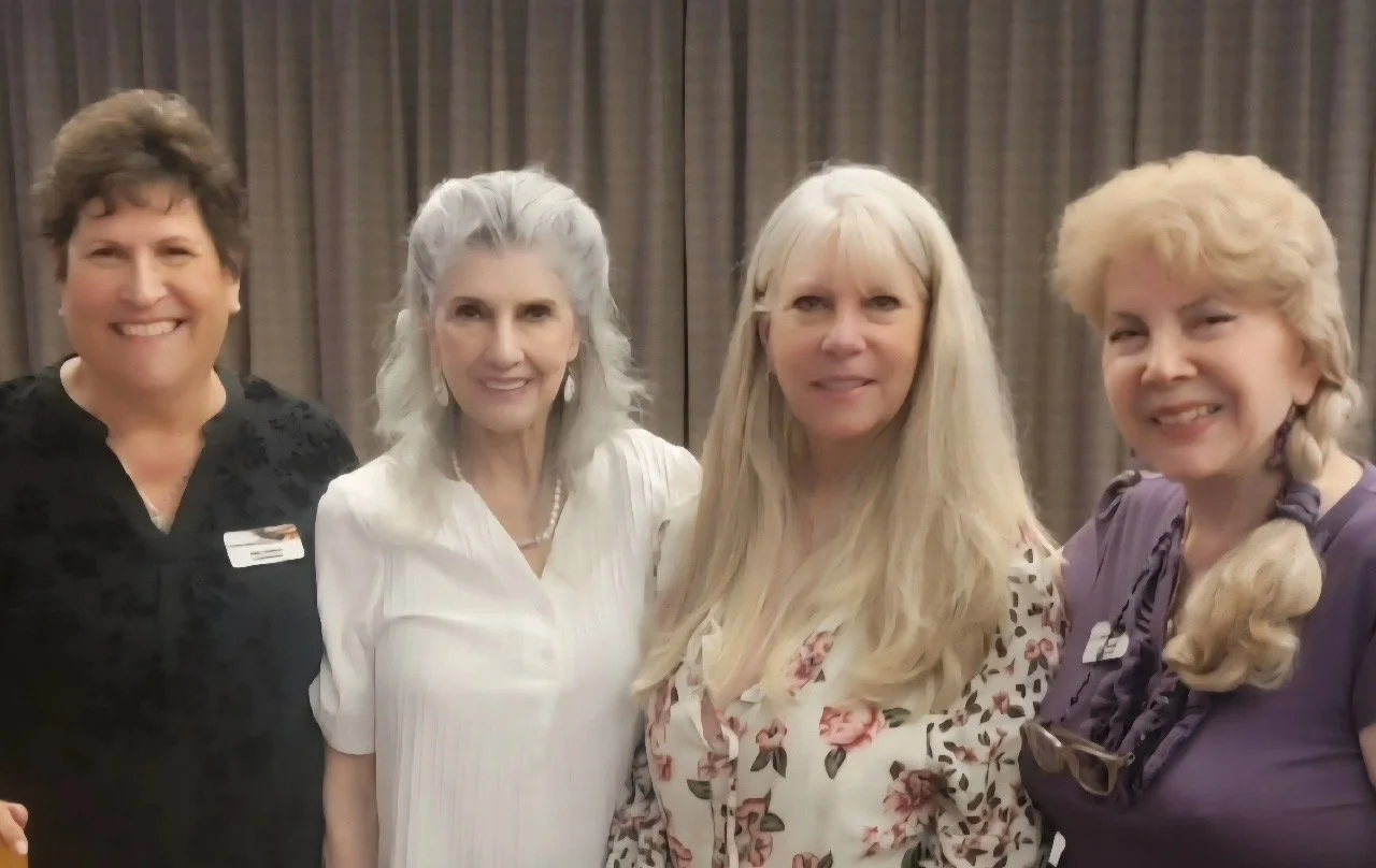 Four women smiling and standing together in front of a brown curtain backdrop.
