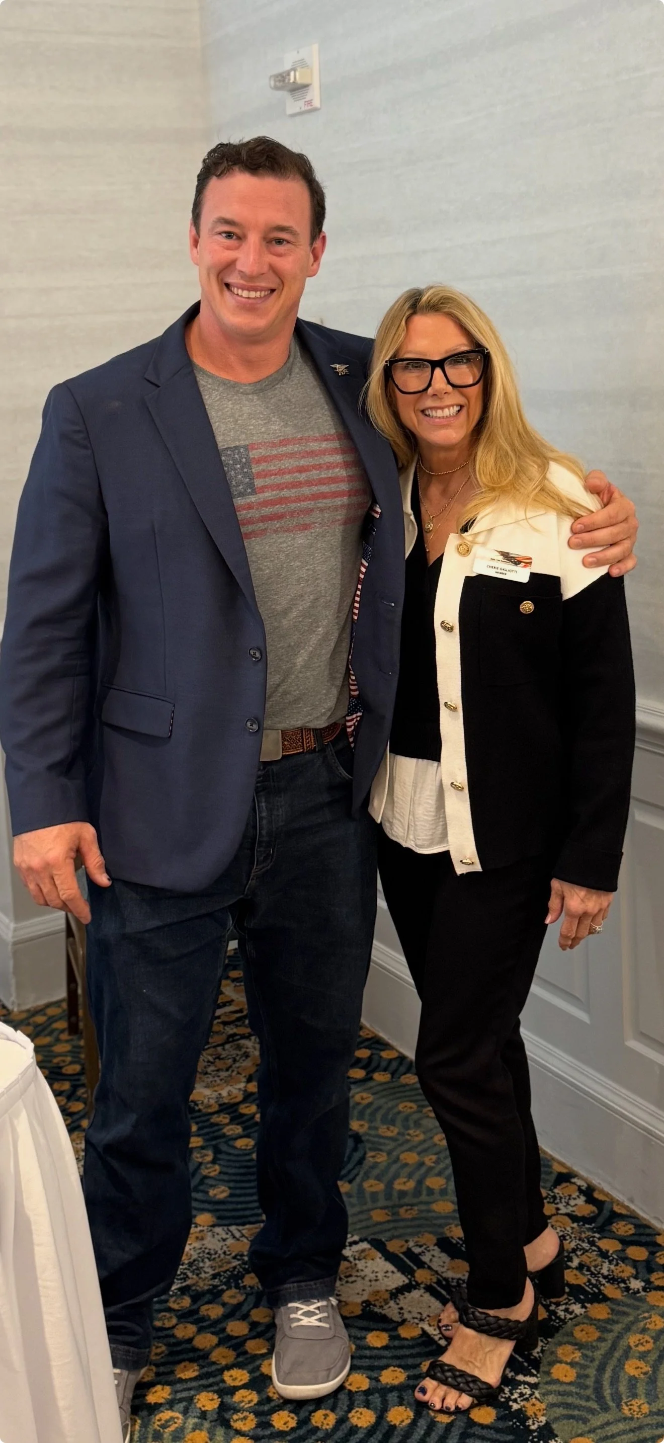 A man and woman standing together indoors, smiling for the camera. The man is wearing a navy blazer, gray T-shirt with an American flag graphic, jeans, and sneakers. The woman is dressed in black pants, a black and white blazer, and black heeled sand