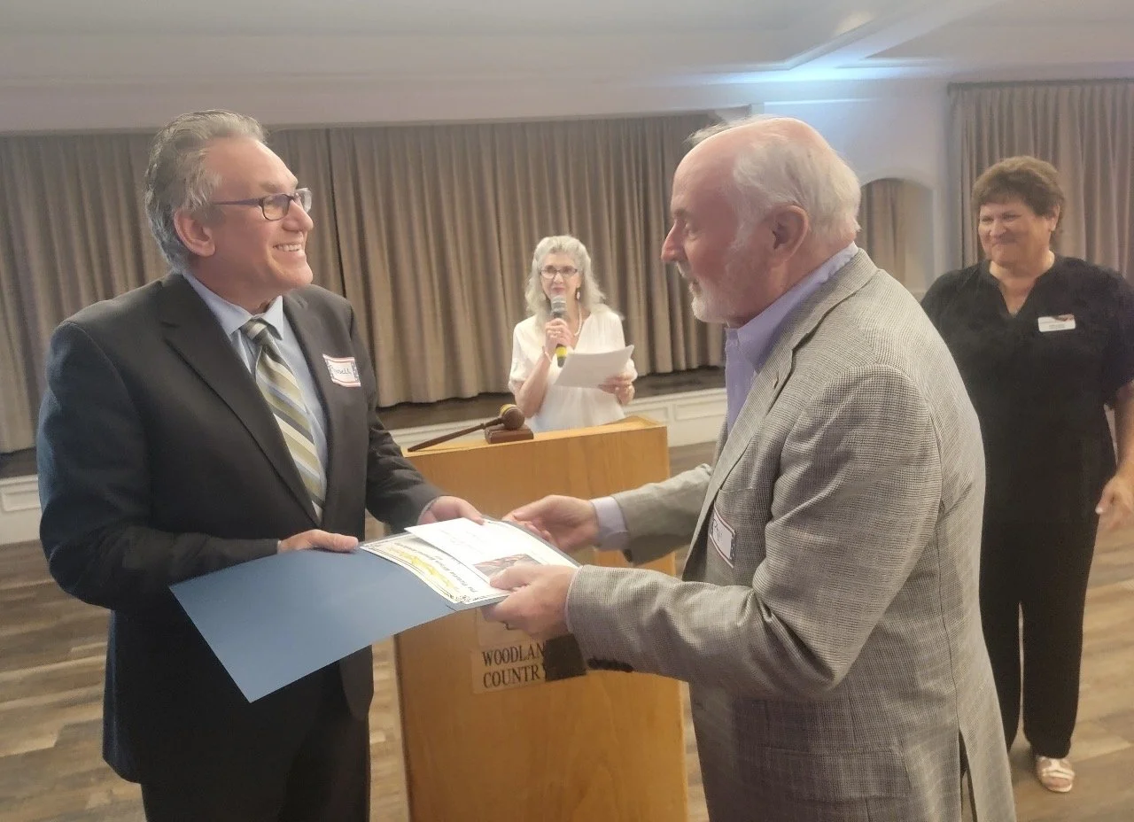 An older man presents a certificate to a younger man at an indoor event, with a woman speaking into a microphone in the background.