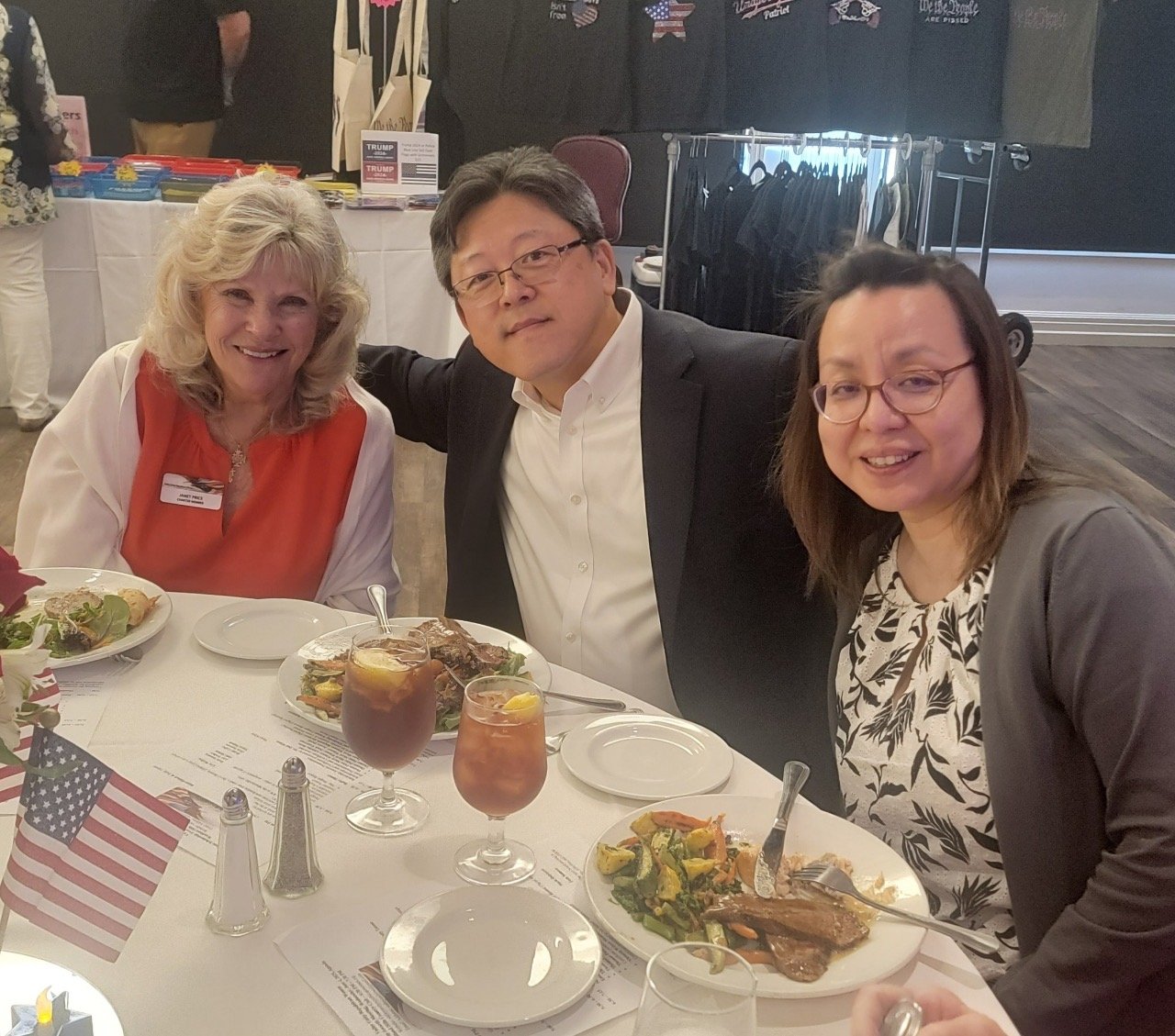 Three people sitting at a table during a meal at a celebration or gathering, with plates of food, drinks, and small American flags on the table.