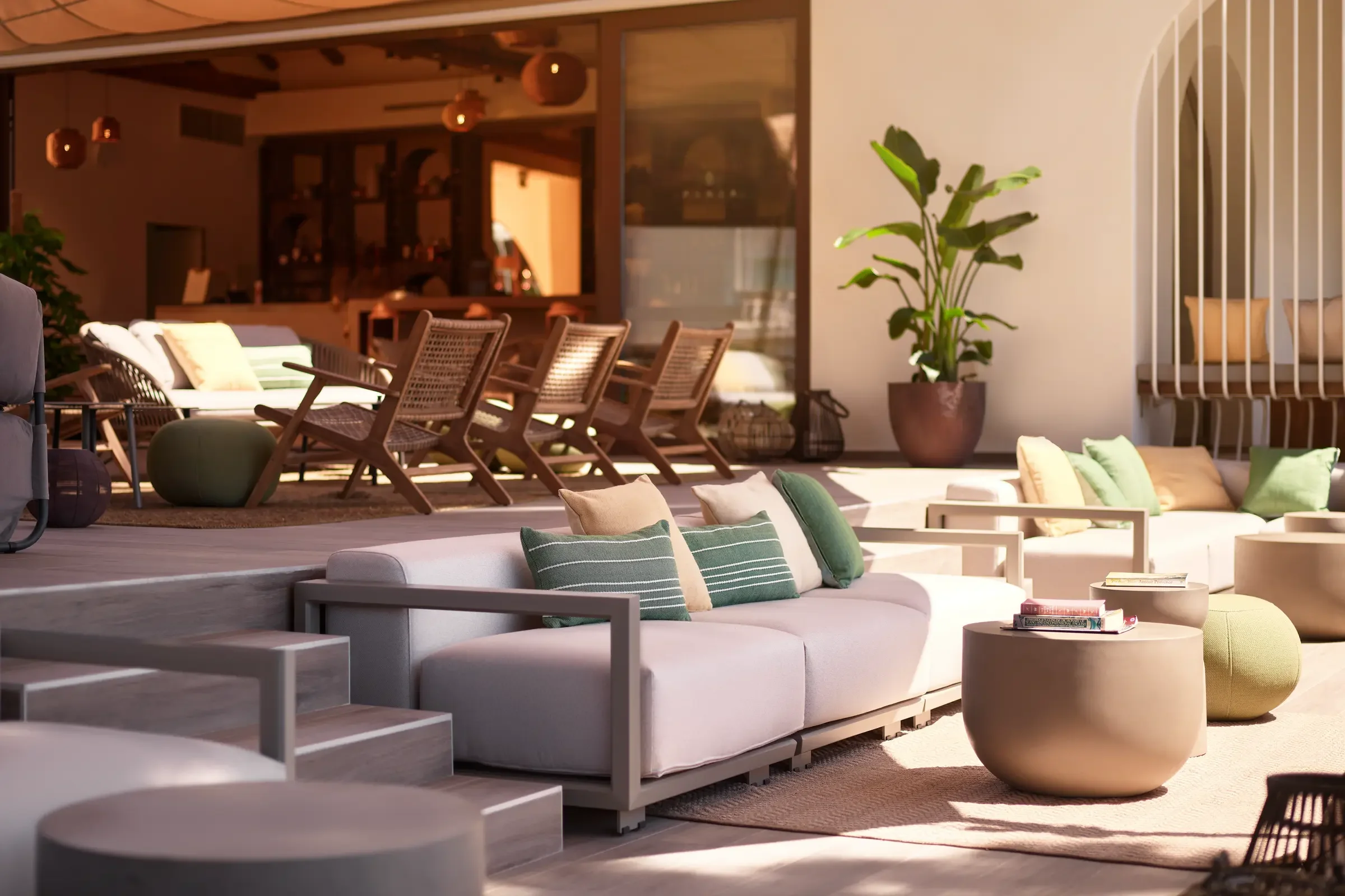 An outdoor lounge area with white sofas, green and beige pillows, round coffee tables with books, and various chairs and potted plants on a patio with sunlight.