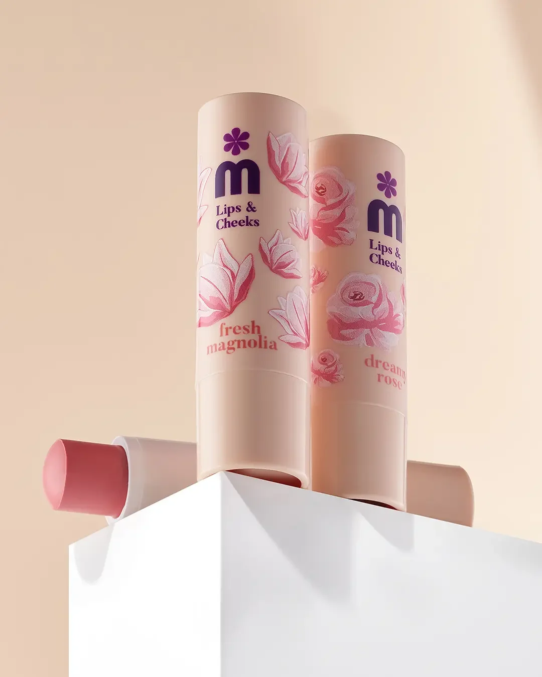 Two cylindrical bottles of M lips and cheeks lip balm on a white surface, one labeled 'fresh magnolia' with pink floral illustrations and the other labeled 'dreamy rose' with pink flowers, against a beige background.