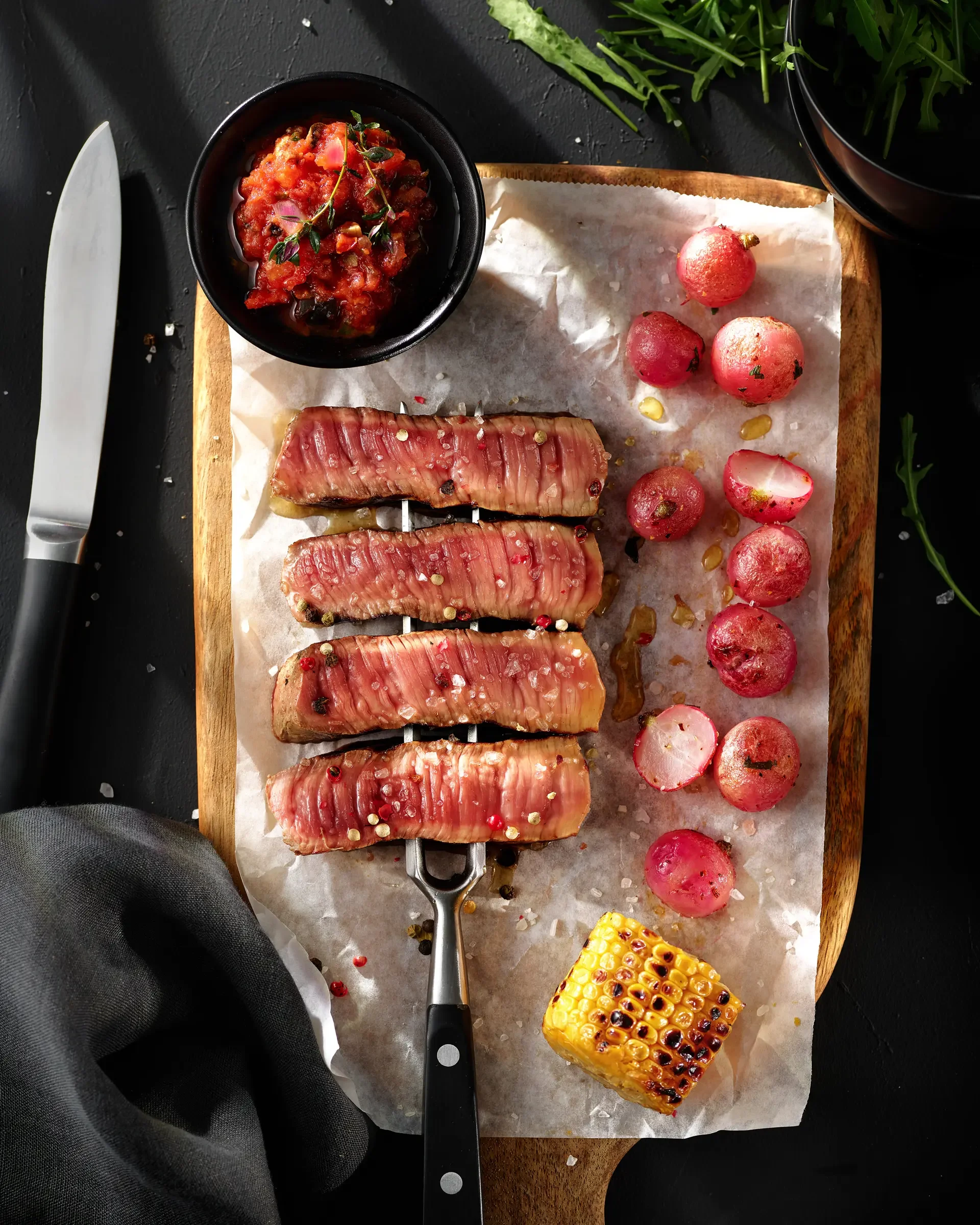 Grilled beef or steak slices topped with coarse salt, served alongside radishes, a small bowl of tomato-based salsa, and a piece of grilled corn on the cob on a wooden cutting board.