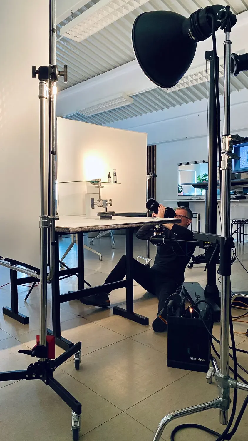 Photographer sitting on the floor taking a picture of a tabletop object, with professional studio lighting and equipment around in a photography studio.