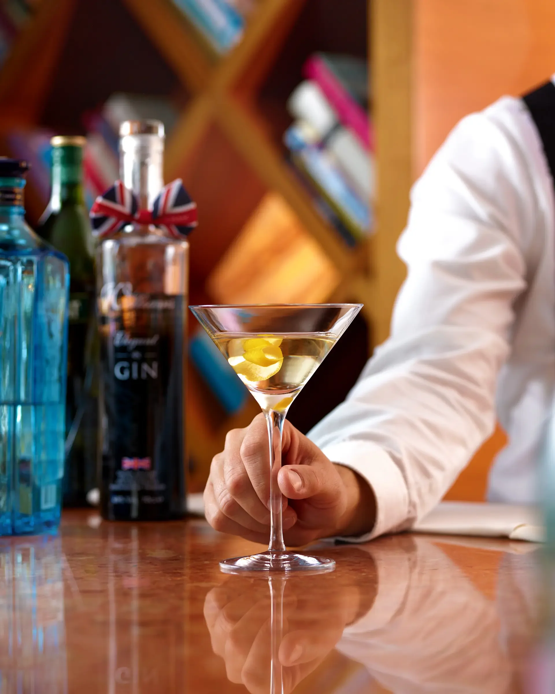 A person in a white shirt holds a martini glass with a lemon twist garnish on a bar counter. Behind, there are bottles of alcohol, including gin, and colorful blurred background decor.