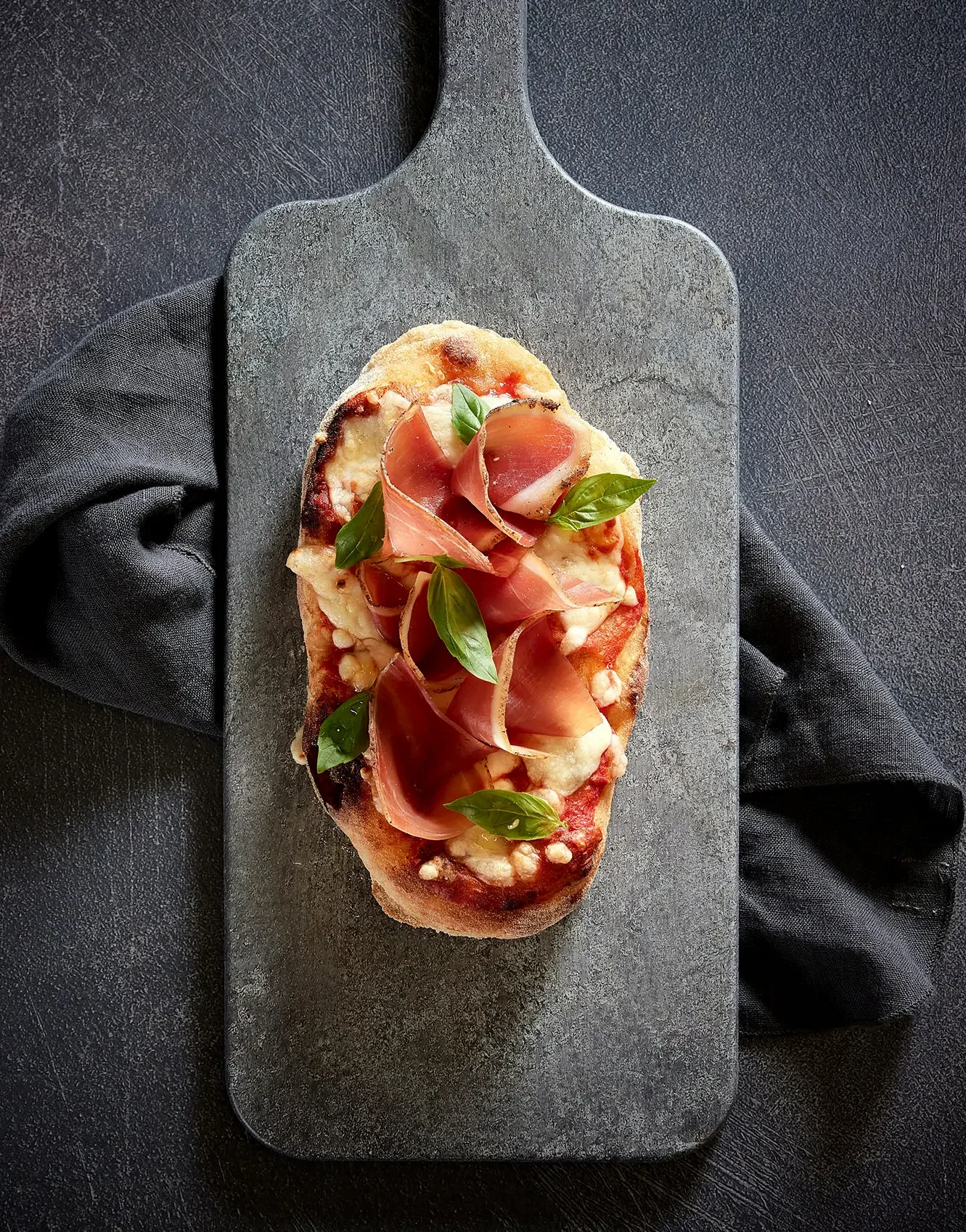 A pizza topped with prosciutto, basil leaves, tomato sauce, and cheese on a black serving board with a black cloth underneath.