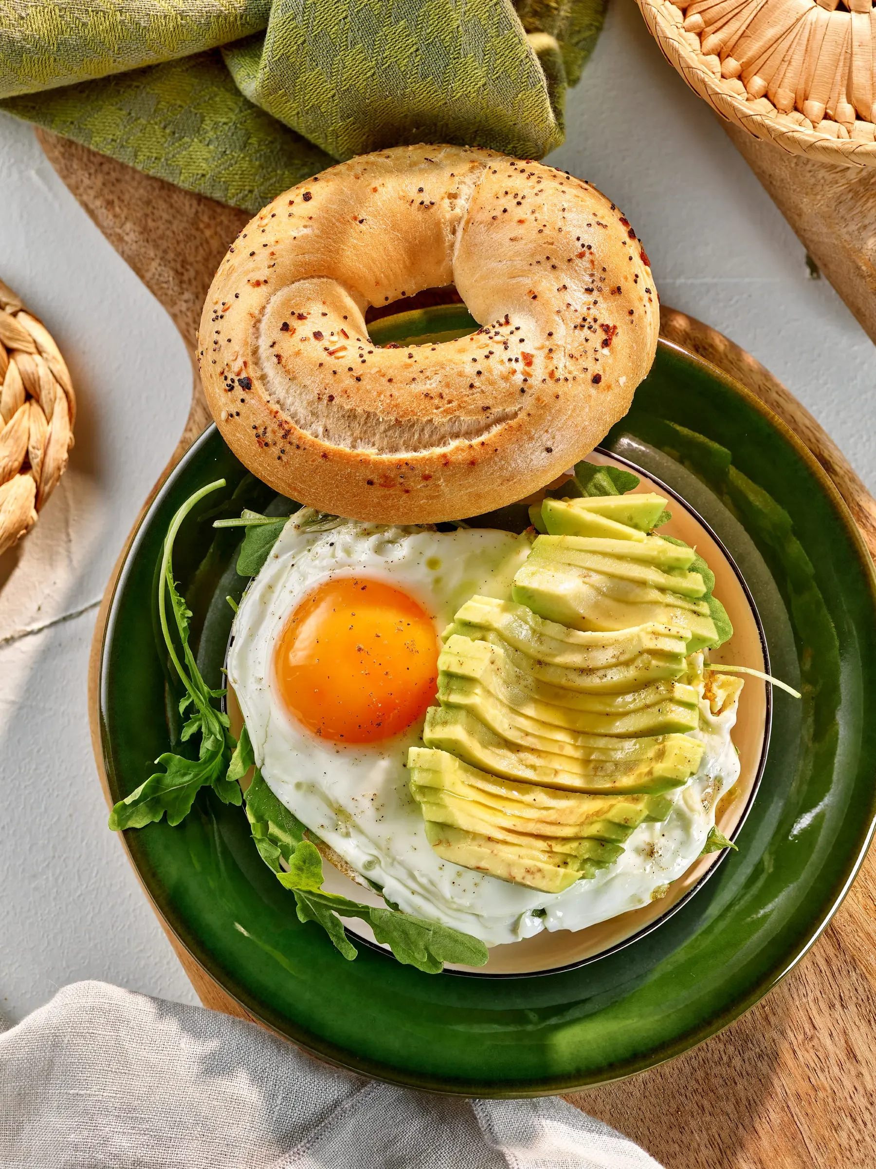 A plate of breakfast food with a sunny side up egg, sliced avocado, and arugula, topped with black pepper, and a bagel with seasoning on top placed on a green plate.
