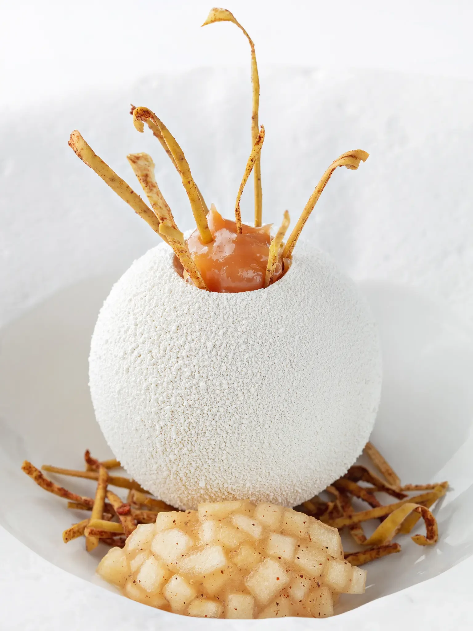 A white, textured sphere resembling an egg, with a caramel center and ginseng roots protruding from the top, surrounded by hash browns and thin crispy strips.