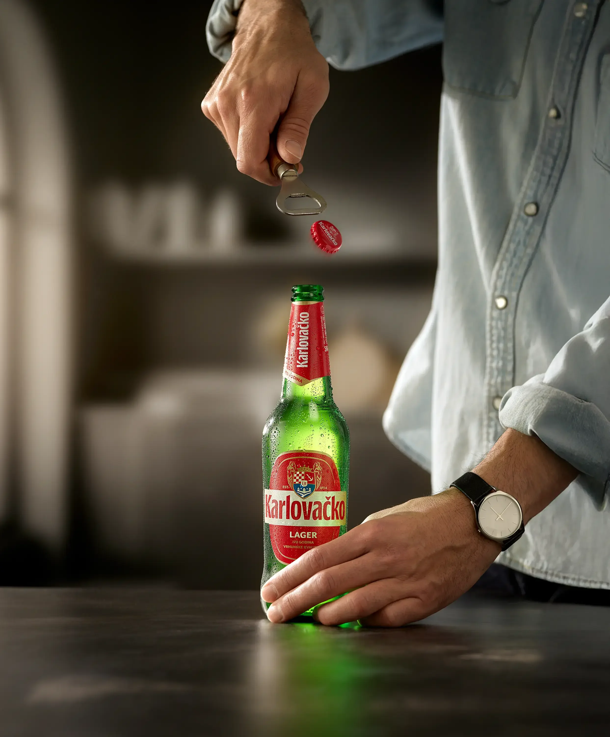 Person opening a green bottle labeled 'Karlovačko Lager' with a bottle opener, it is resting on a dark surface.