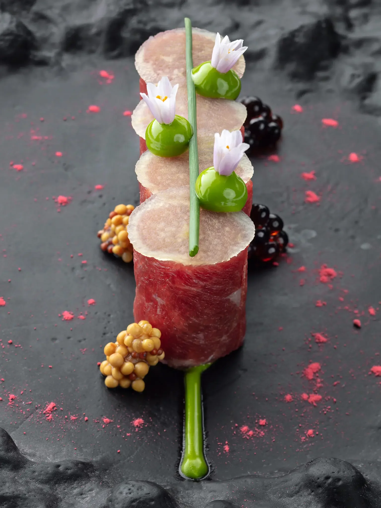 Artistic smoked meat roll topped with thin slices of radish, green spheres, small white flowers, and a green chive, arranged on a black surface with scattered pink and black edible decorations.