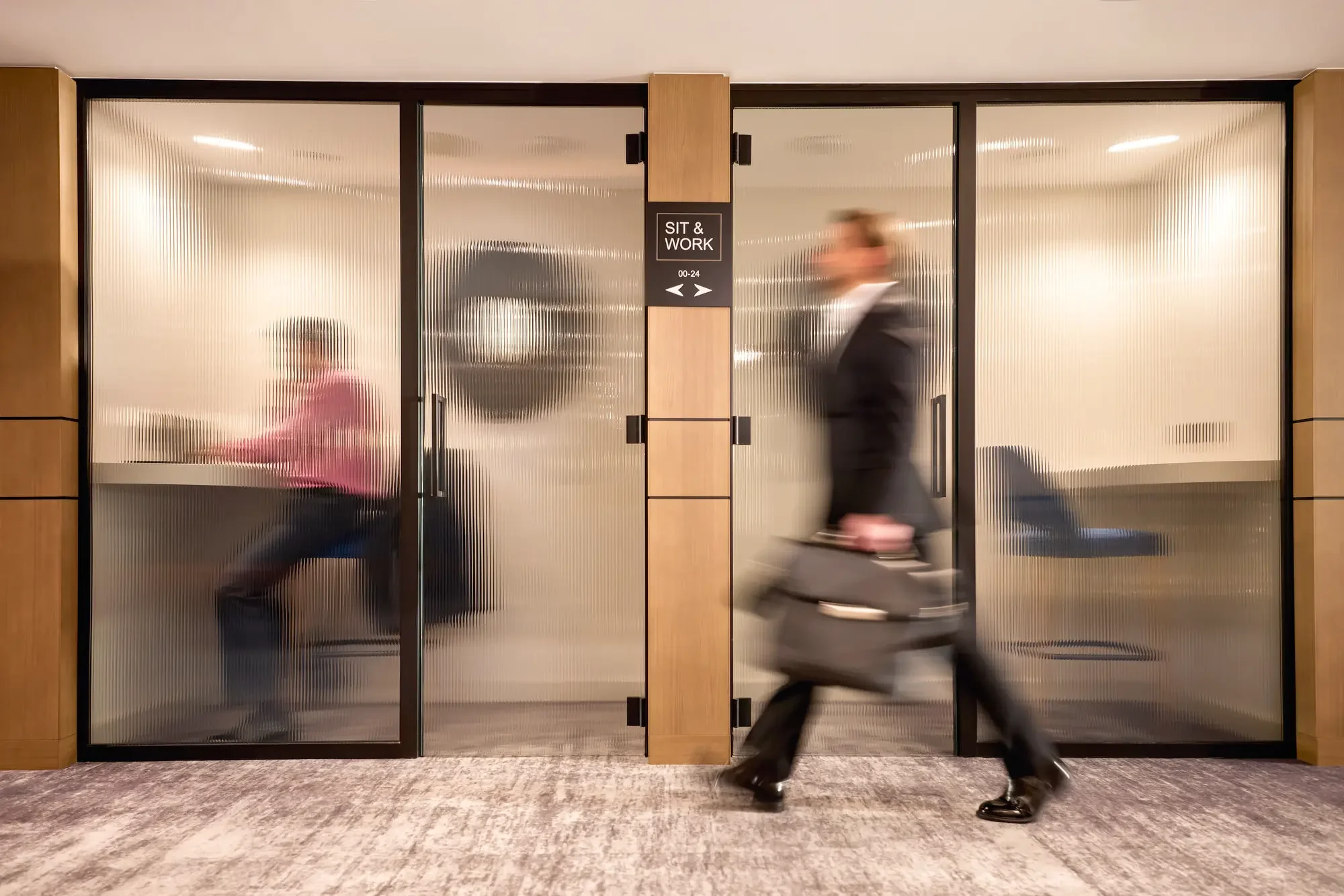 Office interior with glass doors labeled 'Sit & Work', people walking past, one sitting inside, another walking with a briefcase.
