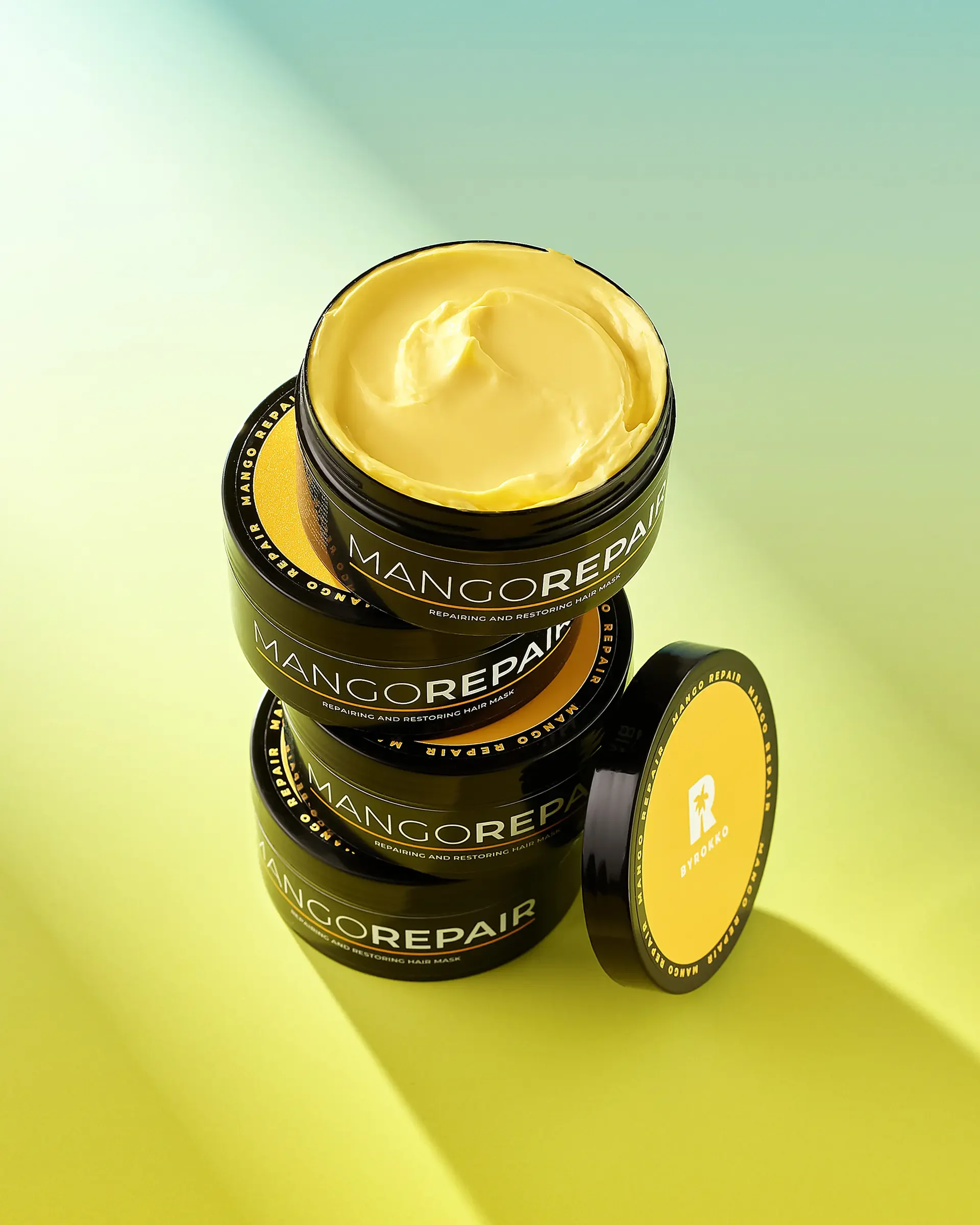 Four stacked black containers of Mango Repair hair mask with yellow labels, one open showing the yellow creamy product, set on a light green background with shadows.
