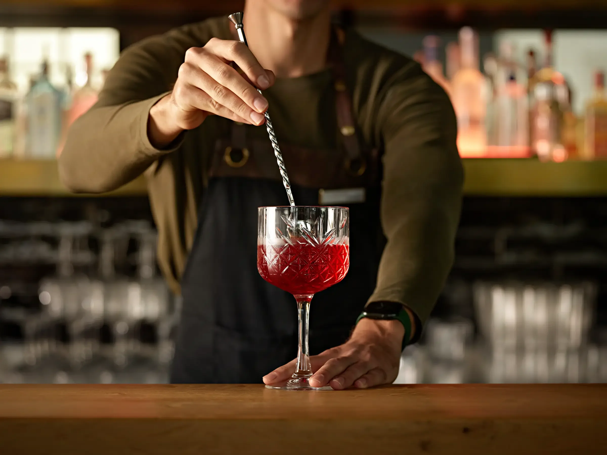 Bartender stirring a red cocktail in a cut-glass with a twisted stir stick behind a wooden bar.