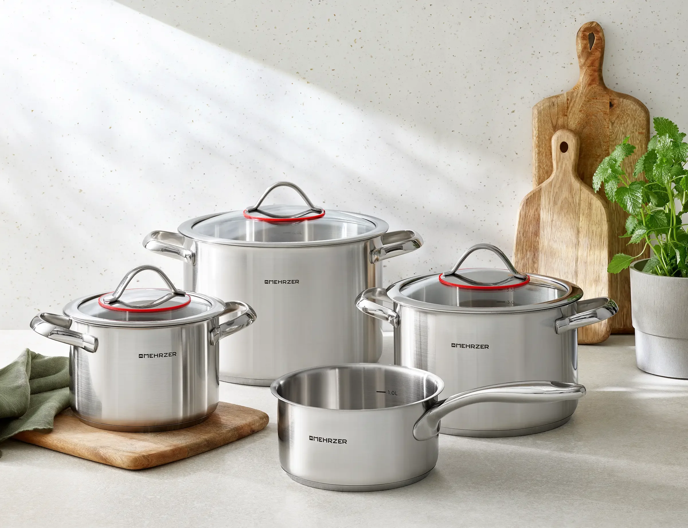 Set of stainless steel cooking pots and pans with glass lids on a kitchen countertop, alongside wooden cutting boards and a potted plant.
