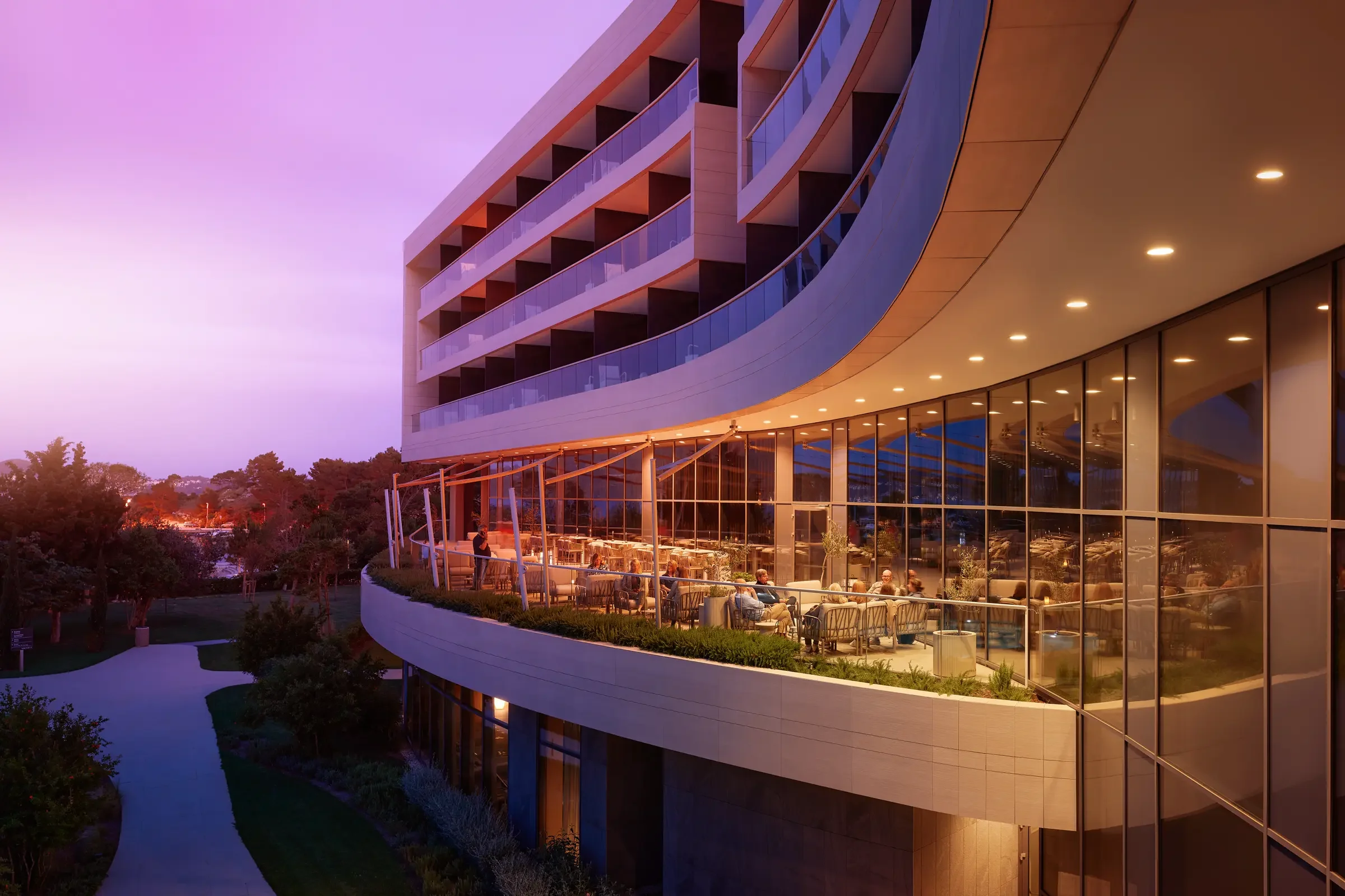 Modern multi-story building with curved glass balconies and an outdoor restaurant on the second floor, illuminated by warm lighting at dusk.