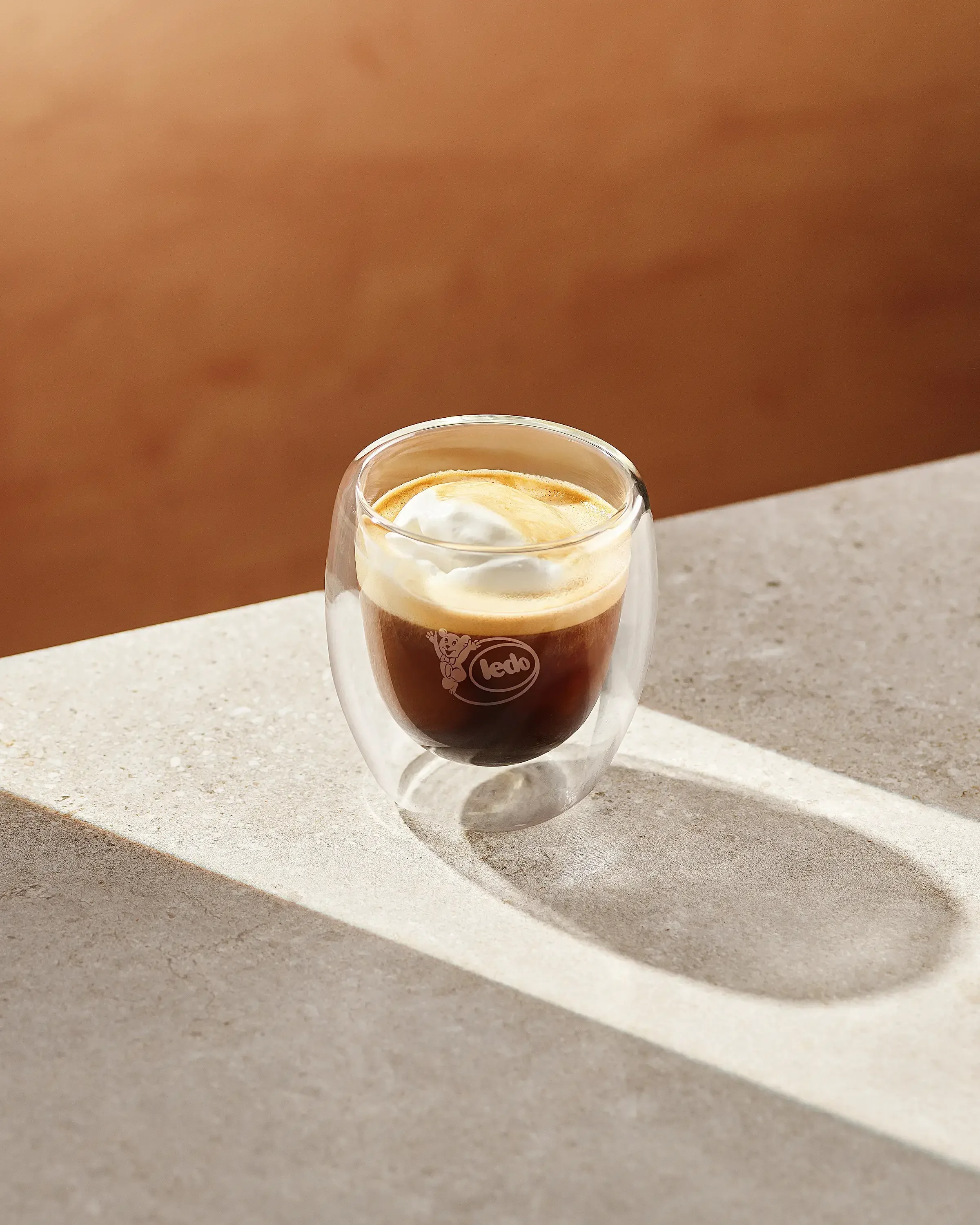A double-walled glass cup of iced coffee topped with a dollop of whipped cream, on a light-colored textured surface with a wooden background.