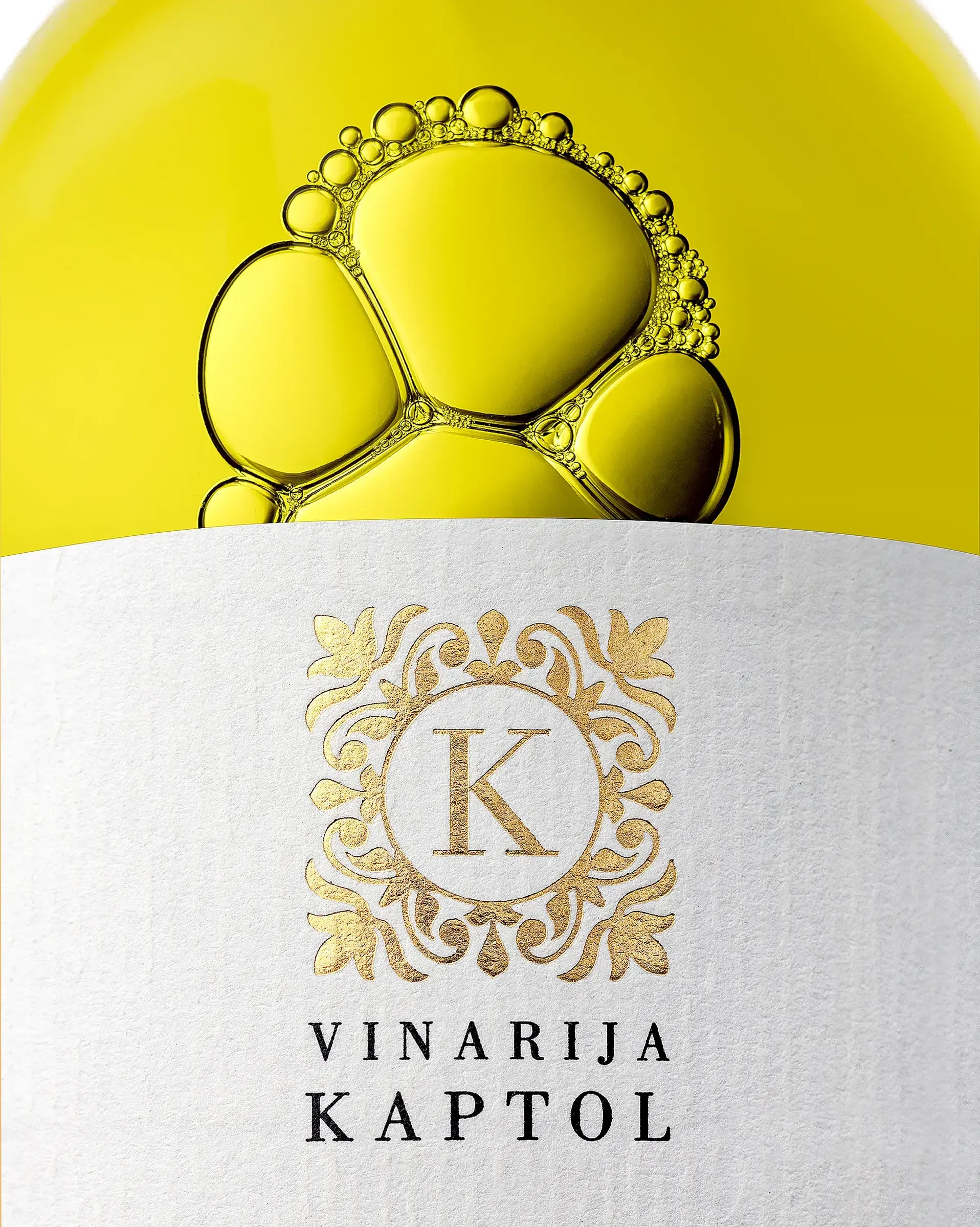 Close-up of a wine bottle label with a gold ornate emblem featuring a letter 'K' and the text 'Vinariја Kaptol' in black, against a white background with a yellow gradient at the top.