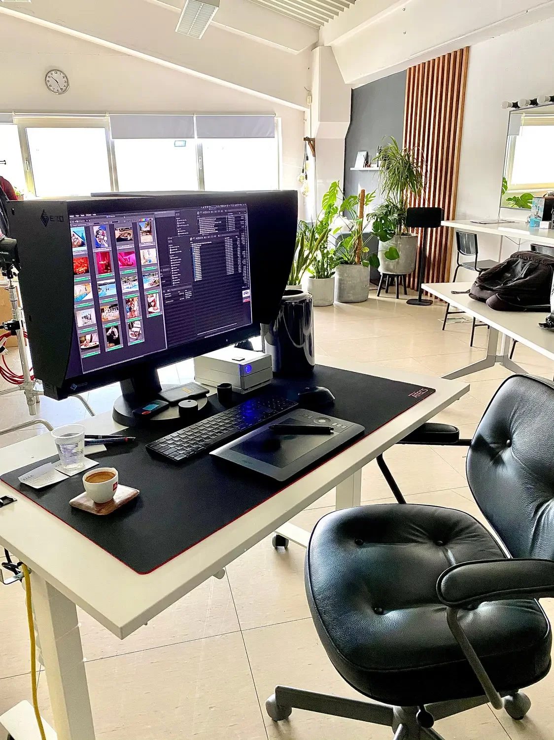 An office workspace featuring a desk with a large monitor, a black office chair, a coffee cup, a graphics tablet, and other accessories. In the background, there are potted plants, a window, and a wall decorated with wooden slats.