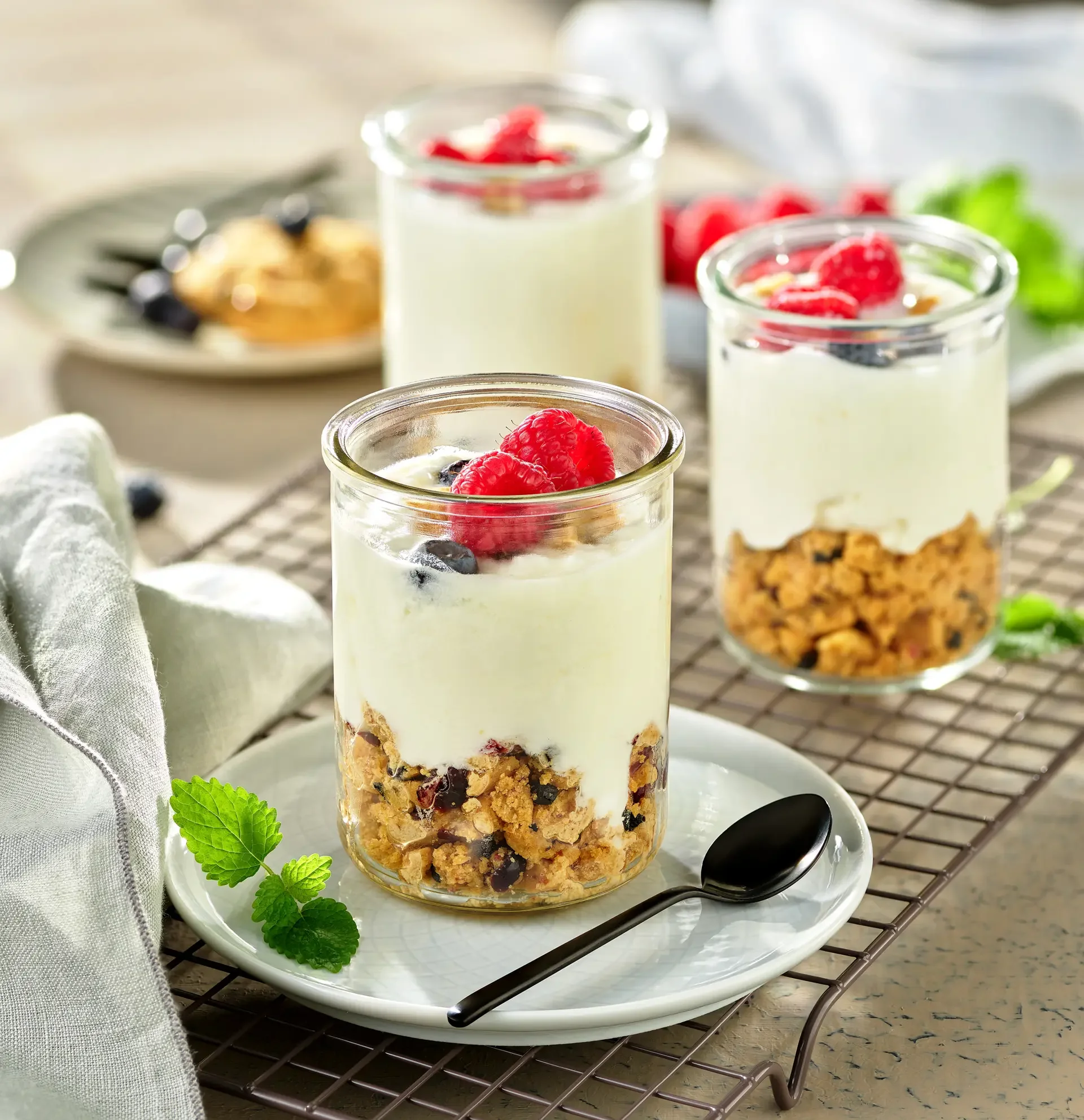 Three glass jars filled with layered dessert, consisting of granola, yogurt, and fresh berries, garnished with mint leaves, on a white plate and a wire cooling rack.