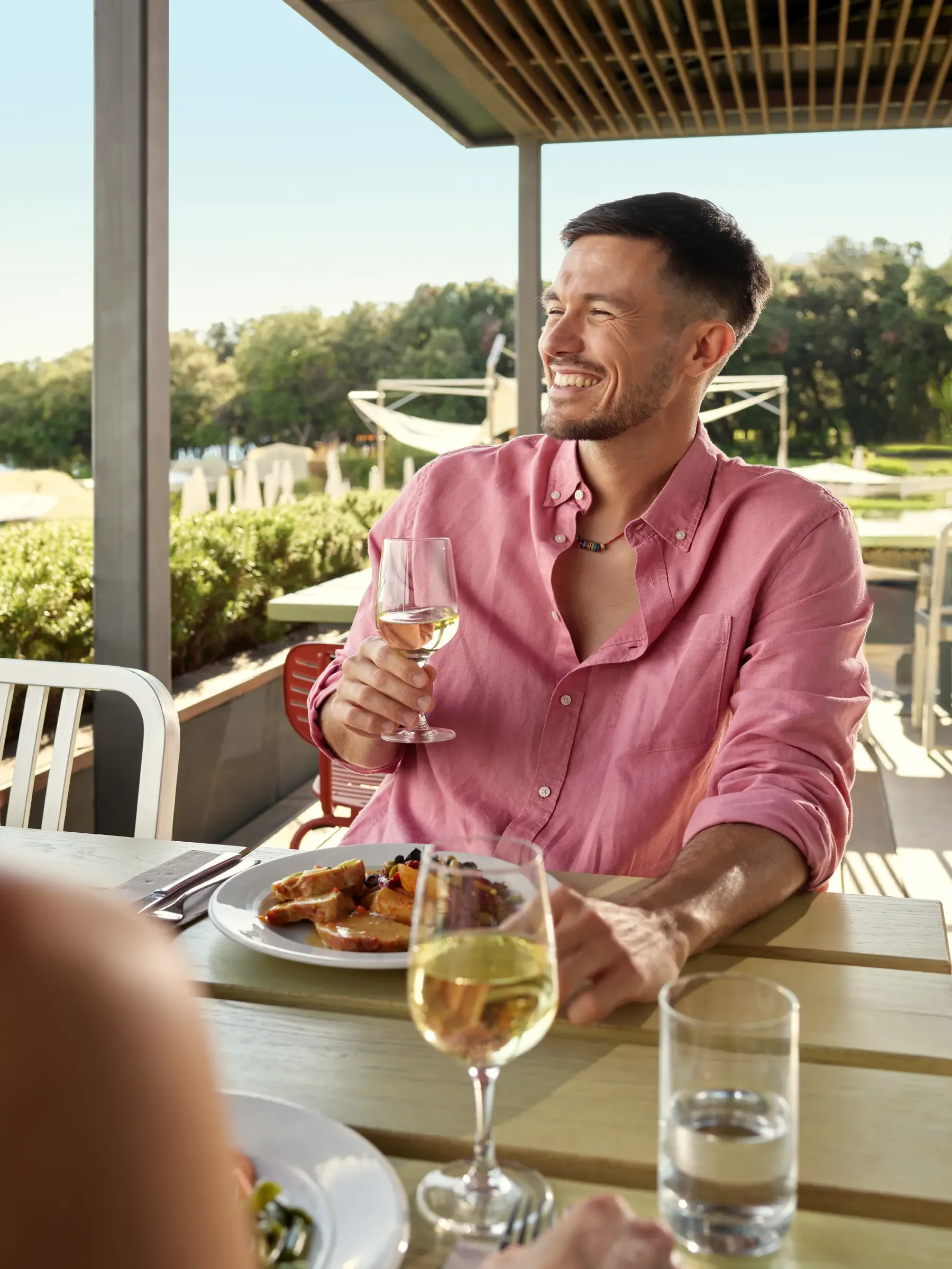 A man in a pink shirt sitting at an outdoor dining table, holding a glass of white wine, smiling. There's a plate of food in front of him, and sunlight with greenery in the background.