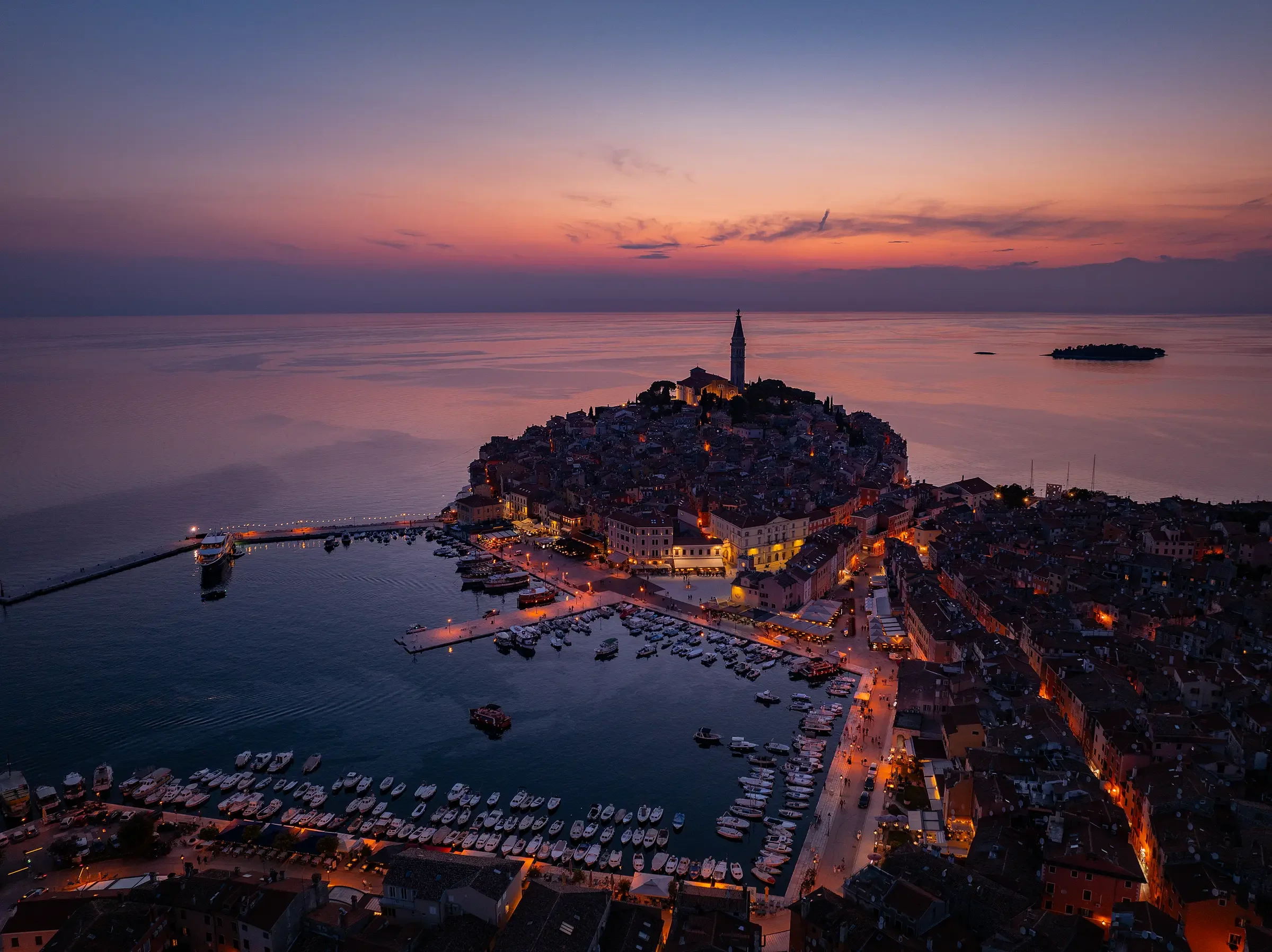 Aerial view of a coastal Italian town at sunset with a hilltop church and town lights reflecting on calm water, boats docked in harbor.