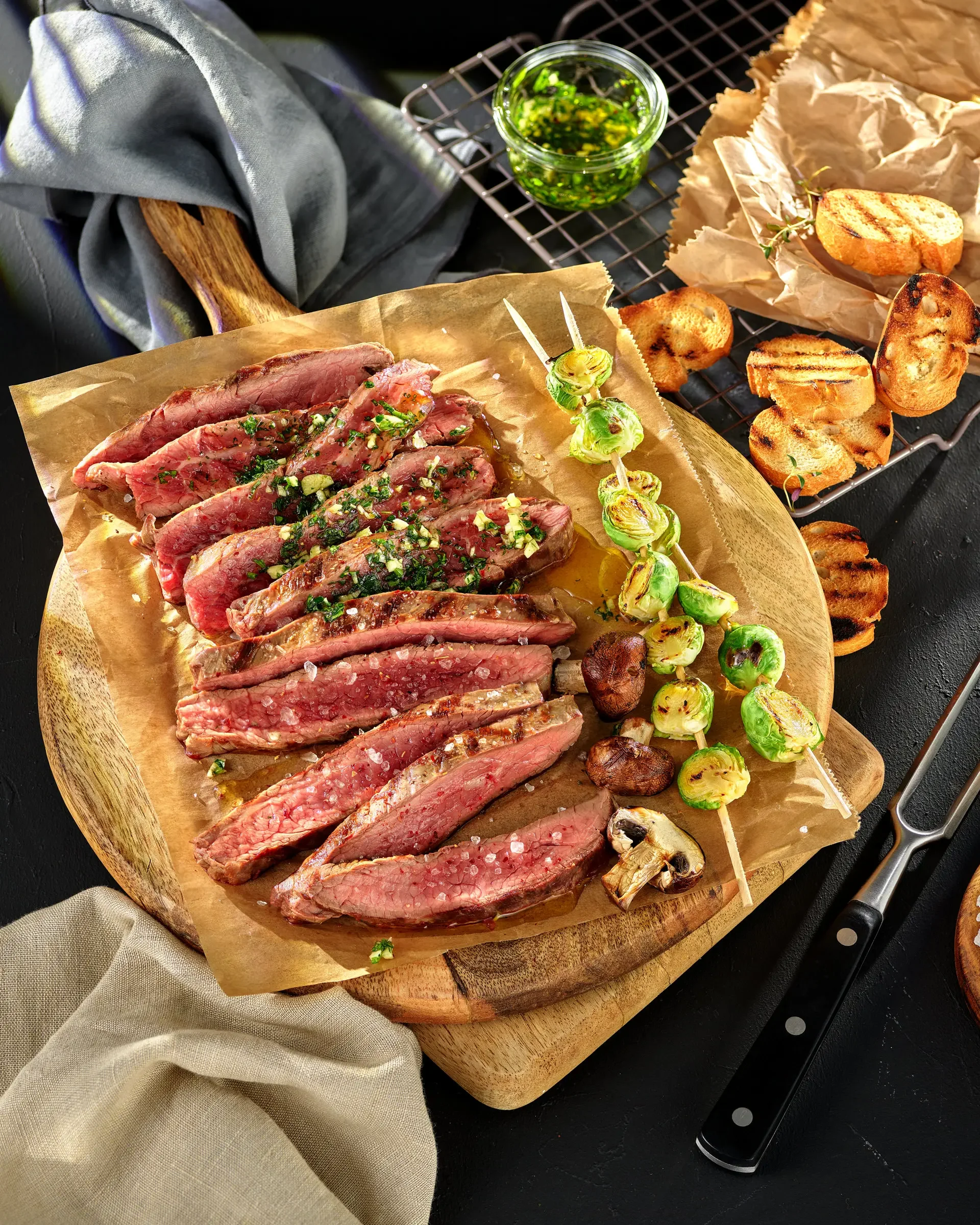 Sliced medium-rare steak garnished with herbs on a wooden serving board, with grilled Brussels sprouts on skewers, and a knife nearby. In the background, a tray of grilled bread, a small bowl of green herb sauce, and crumpled parchment paper.