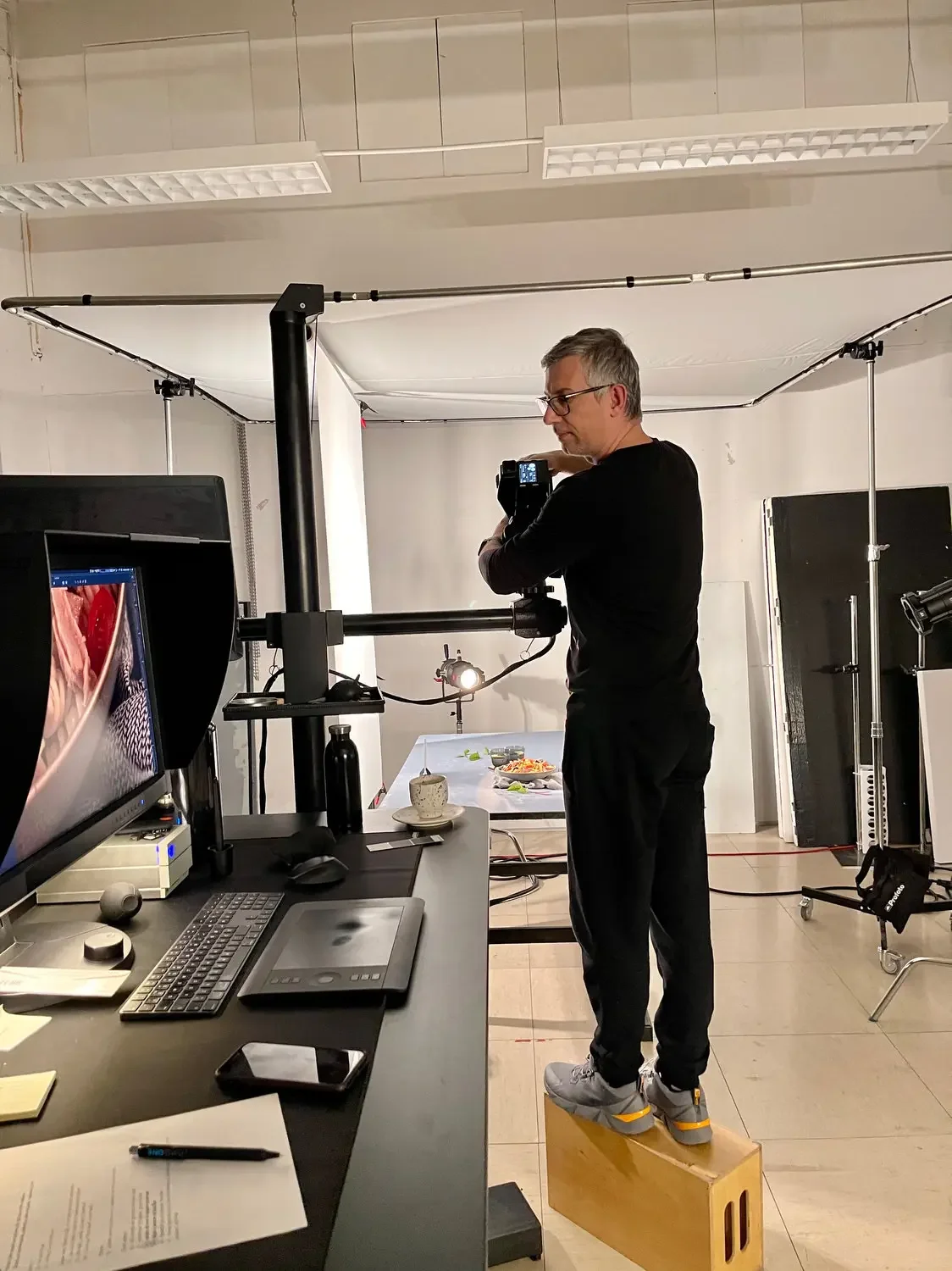 A man standing on a wooden block in a photography studio, adjusting a camera on a tripod, with a backdrop and studio equipment around him.