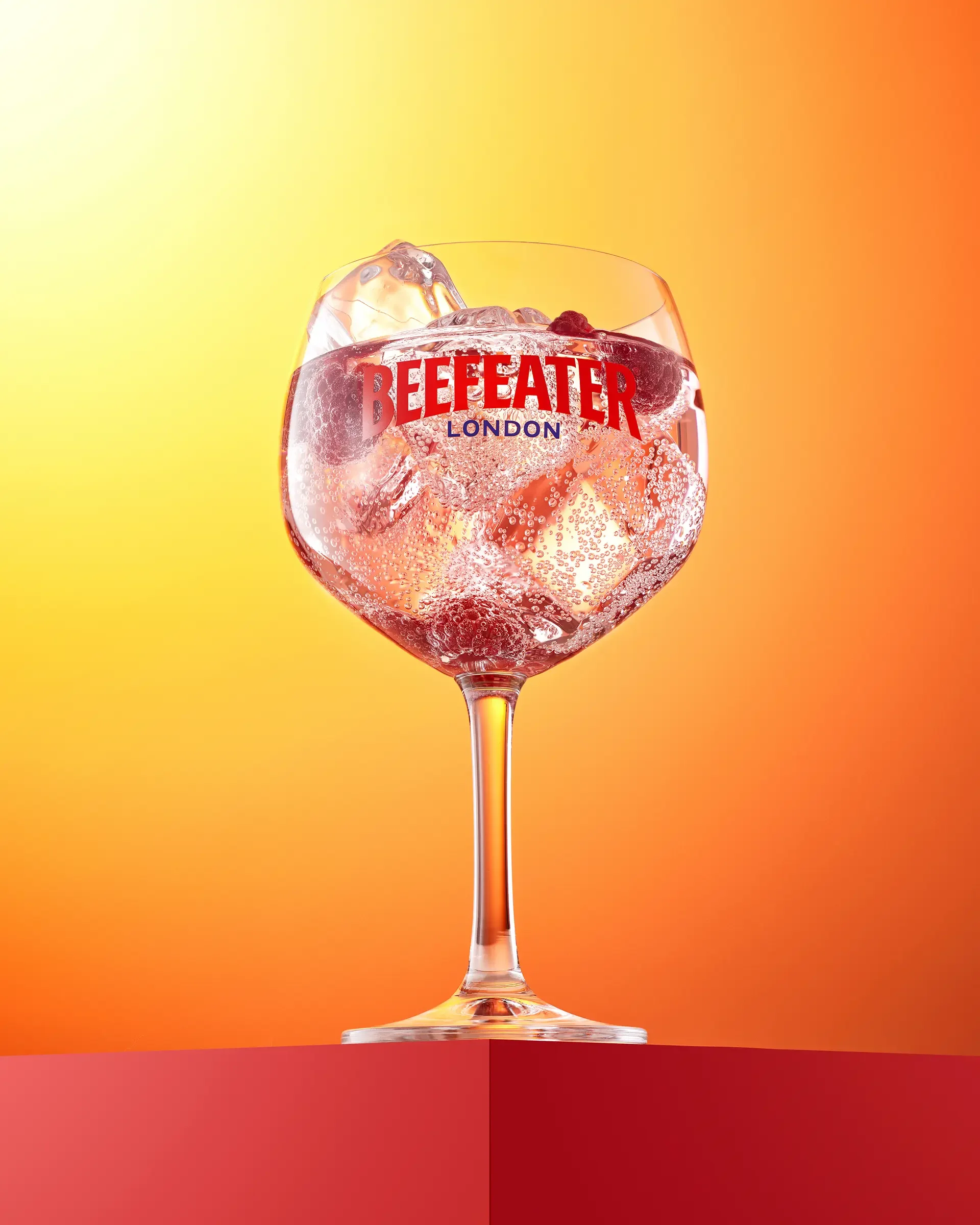 A large wine glass filled with clear soda, ice cubes, and ice cubes with blackberries inside. The glass has the text 'BEEFEATER LONDON' on it, and the background is a gradient of yellow, orange, and red.