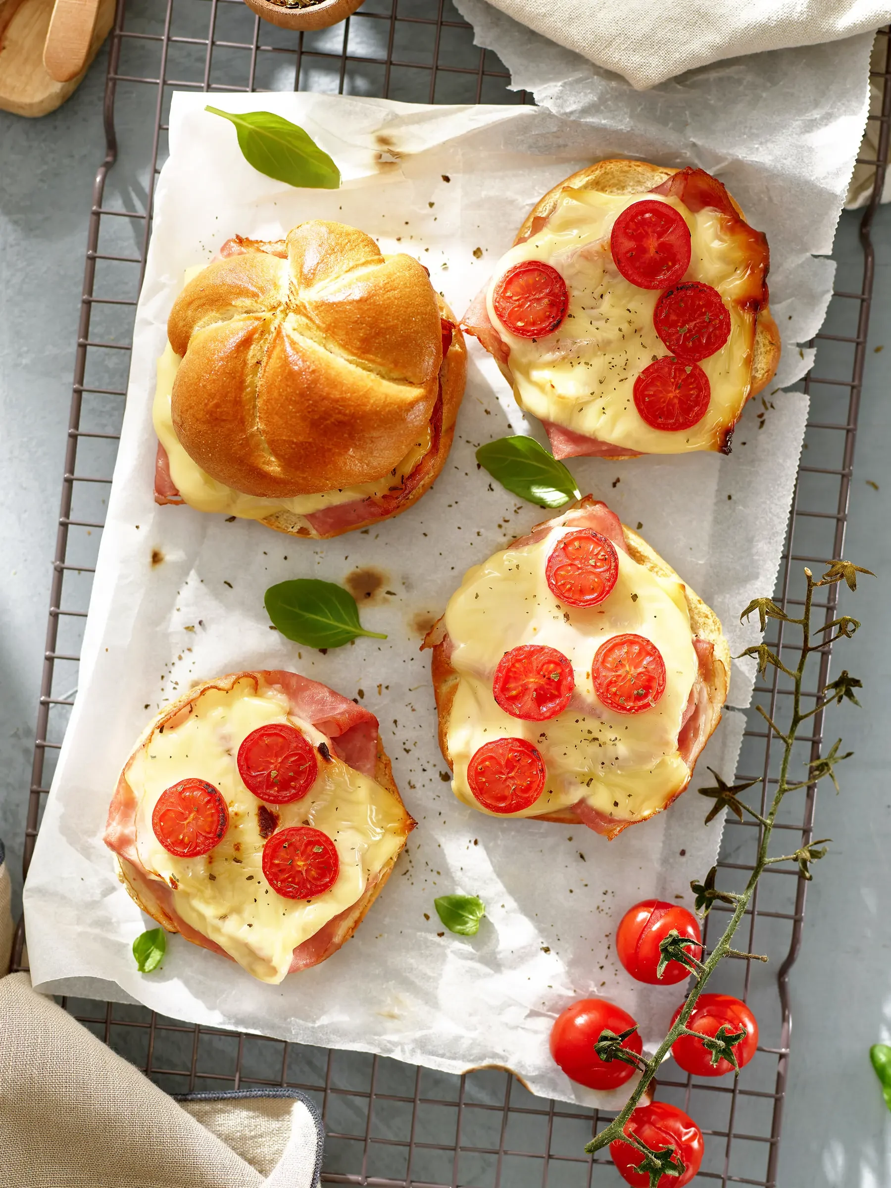 Three open-faced sandwiches with melted cheese, cherry tomatoes, and ham, served on a metal rack lined with parchment paper, garnished with fresh basil leaves and surrounded by cherry tomatoes.