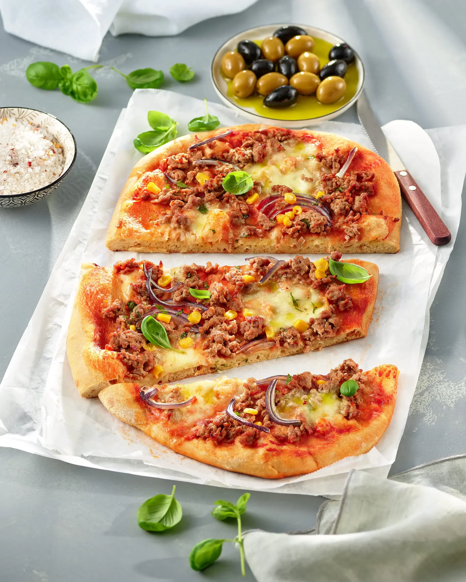 Slices of pizza topped with ground meat, red onions, yellow corn, and fresh basil leaves, on parchment paper with bowls of mixed olives, sea salt, and fresh herbs in the background.