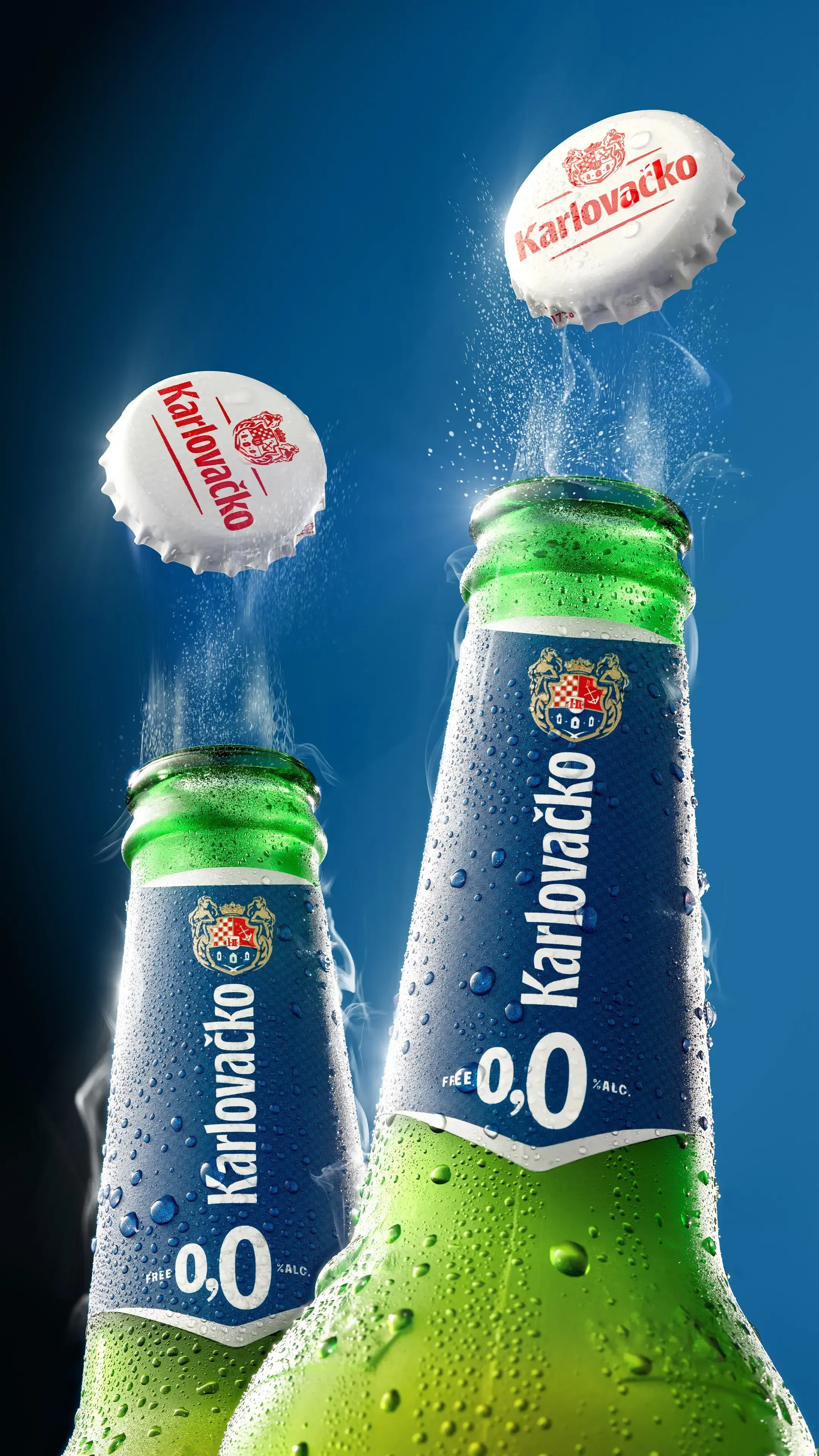 Two Karlovacko beer bottles with condensation, against a blue sky, with caps flying off from the top.