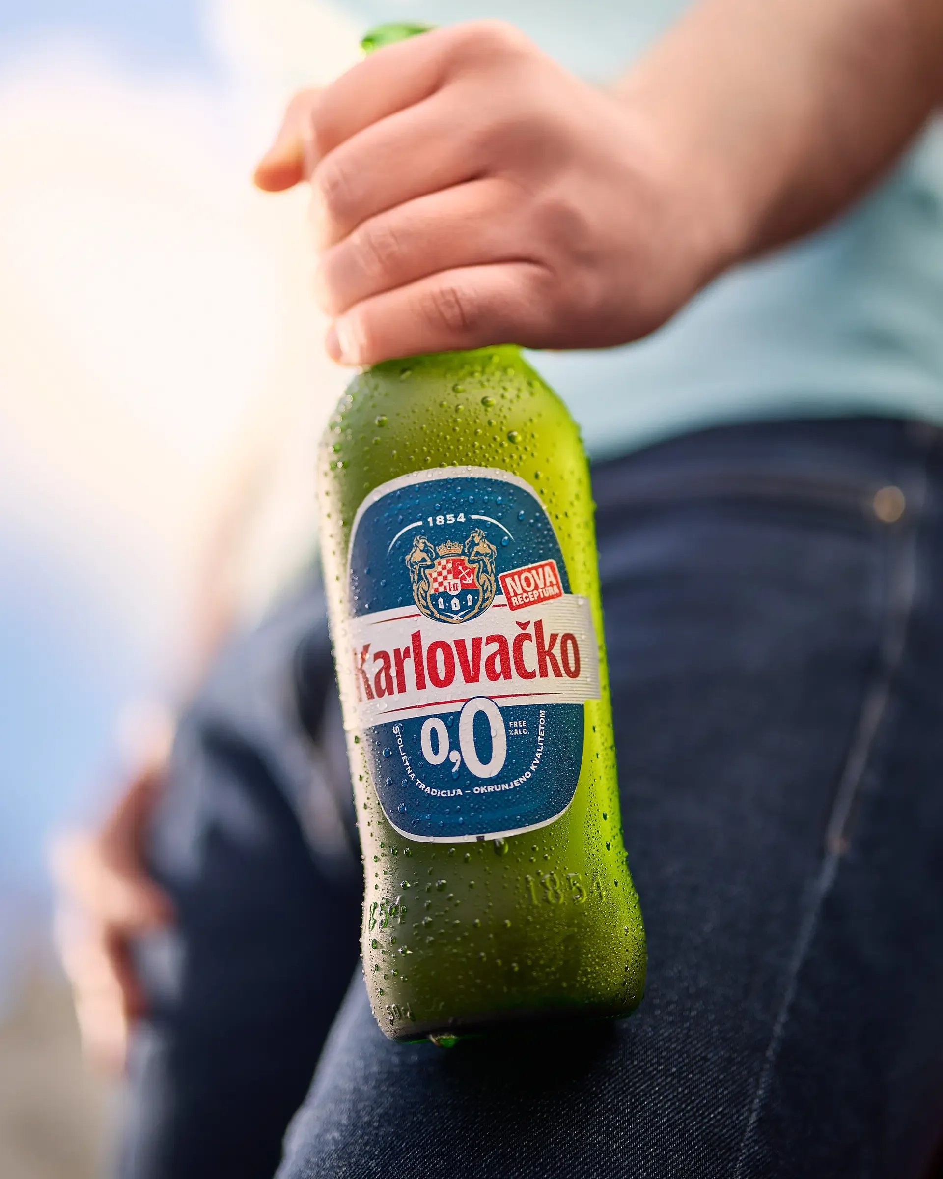 A person holding a chilled Karlovačko 0.0 non-alcoholic beer bottle covered in condensation.