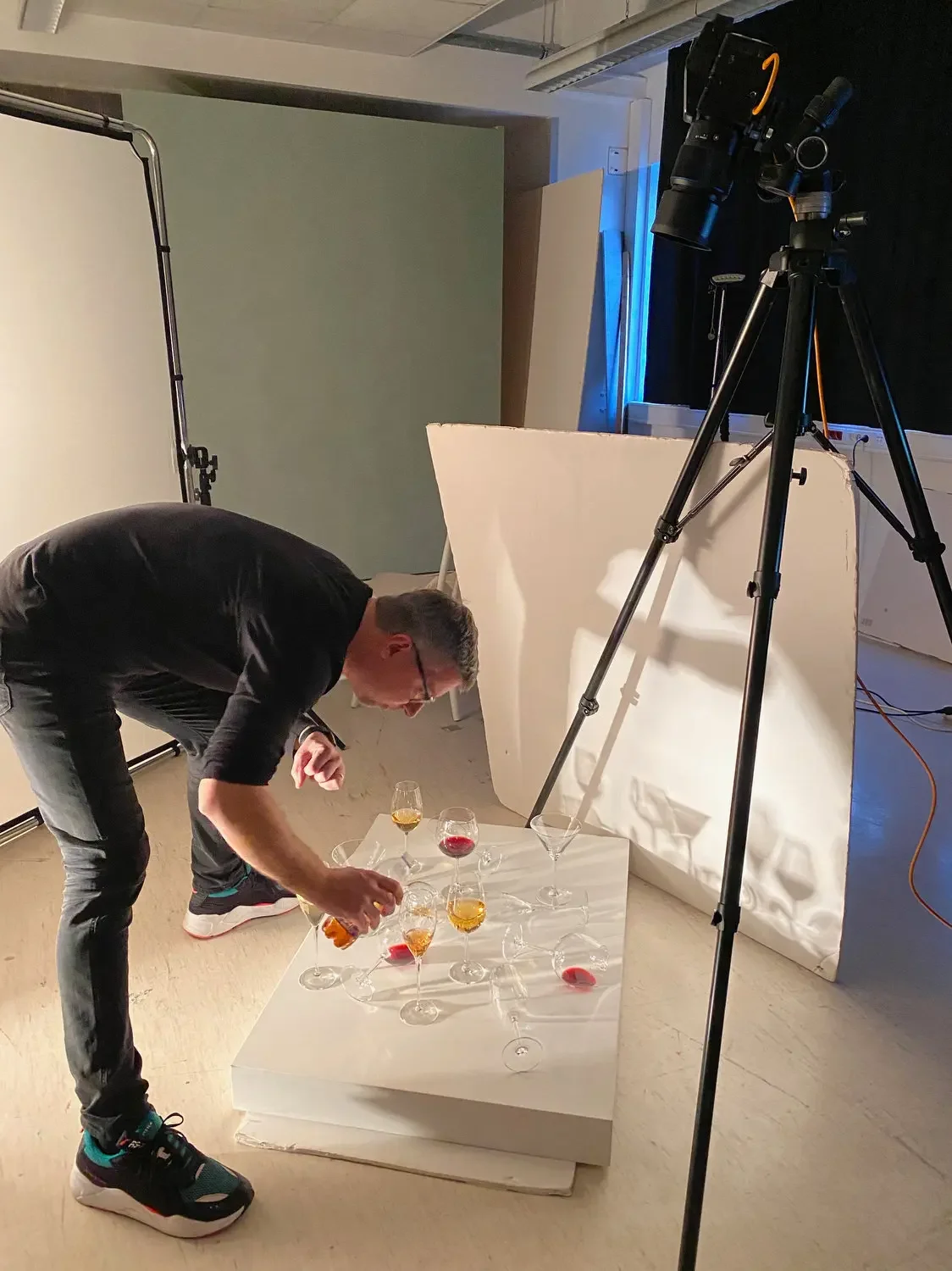 A man arranging glasses with colorful drinks on a white platform in a photography studio, with professional lighting and equipment visible.