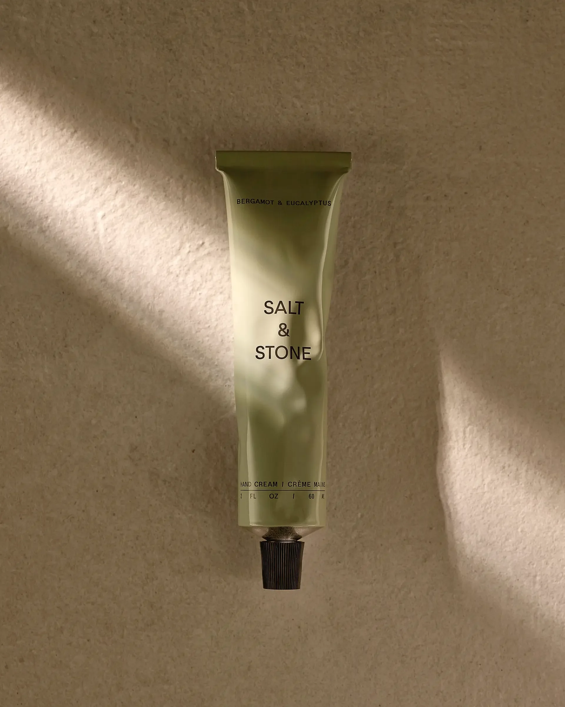 A tube of Bergamot & Eucalyptus scented hand cream labeled 'Salt & Stone' on a beige surface with textured fabric. Sunlight casts shadows on the surface.