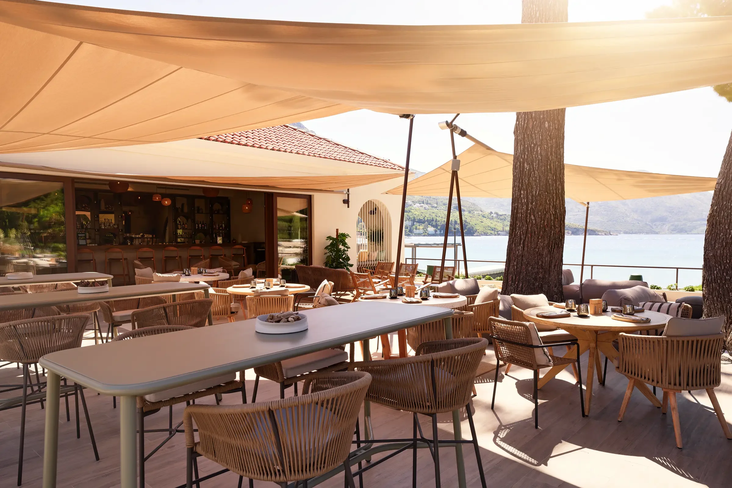 An outdoor restaurant patio overlooking a body of water, with various tables and chairs under large beige umbrellas and shaded areas, surrounded by tall trees.