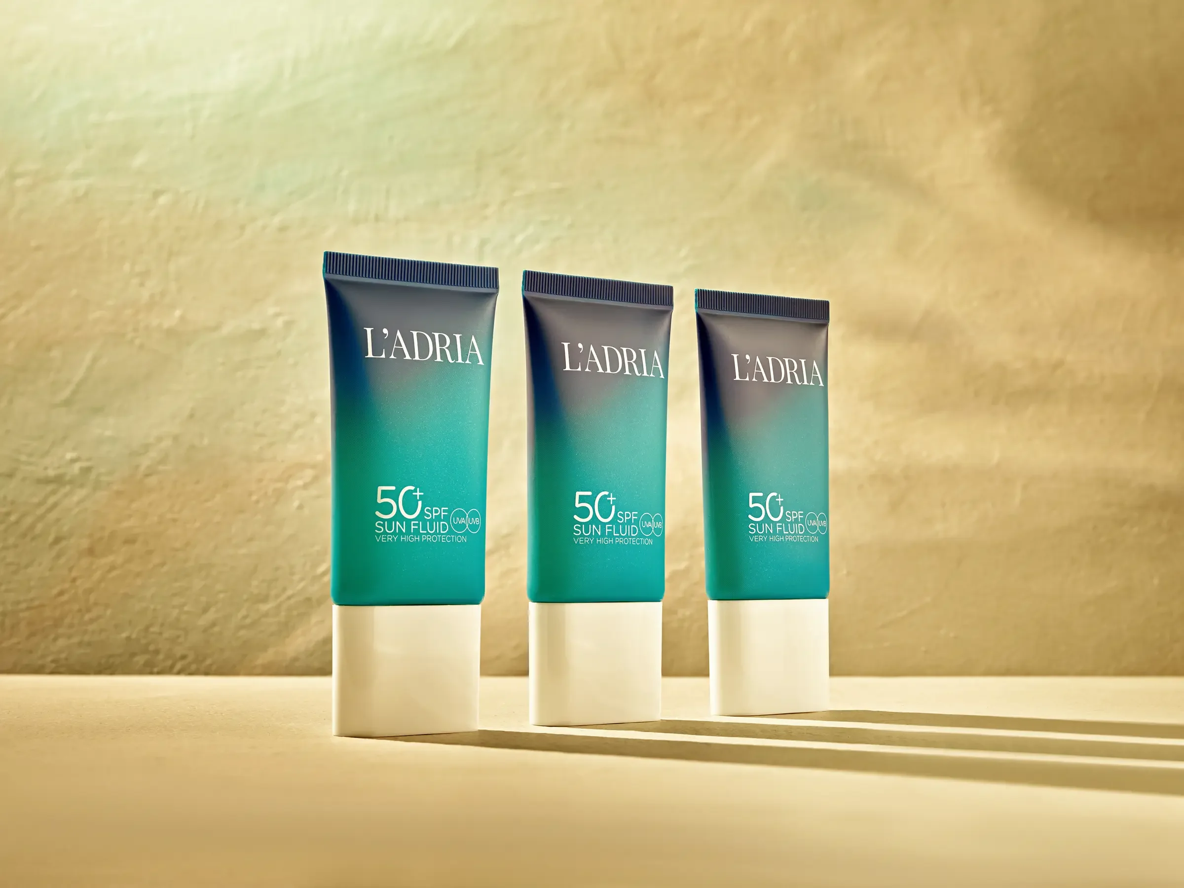 Three tubes of L'Adria 50+ SPF sun fluid standing on a surface with shadows cast behind them, against a textured beige background.