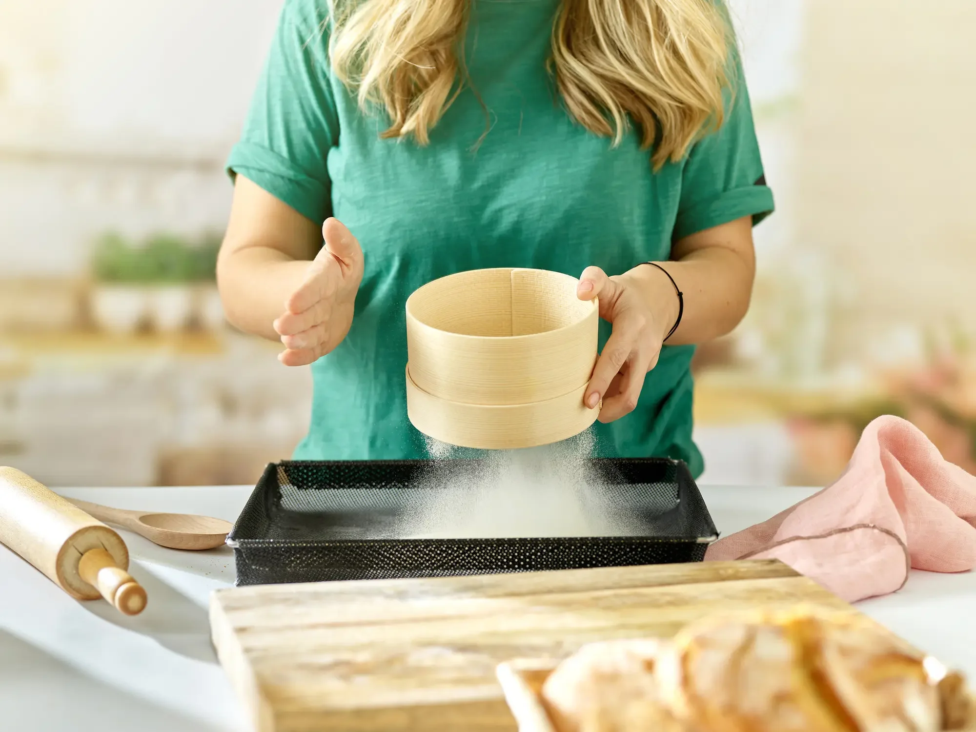 A woman sifting powdered sugar into a baking pan in a kitchen with a rolling pin and wooden spoon nearby.