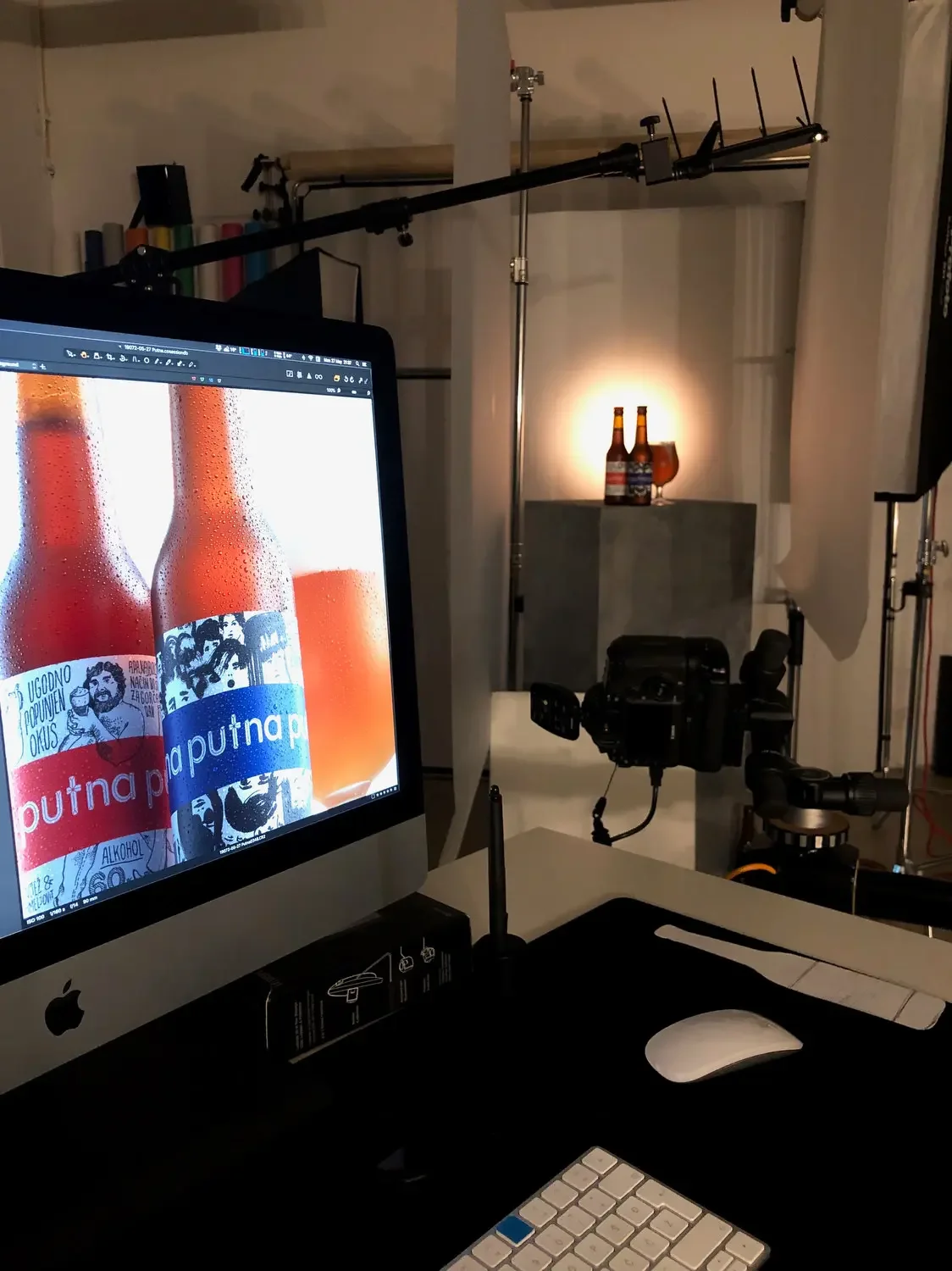 Studio setup with an Apple iMac computer displaying beer bottles with colorful labels, surrounded by photography and video equipment including a camera, tripod, and lighting, with additional beer bottles and a wine glass on a side table in the backgr