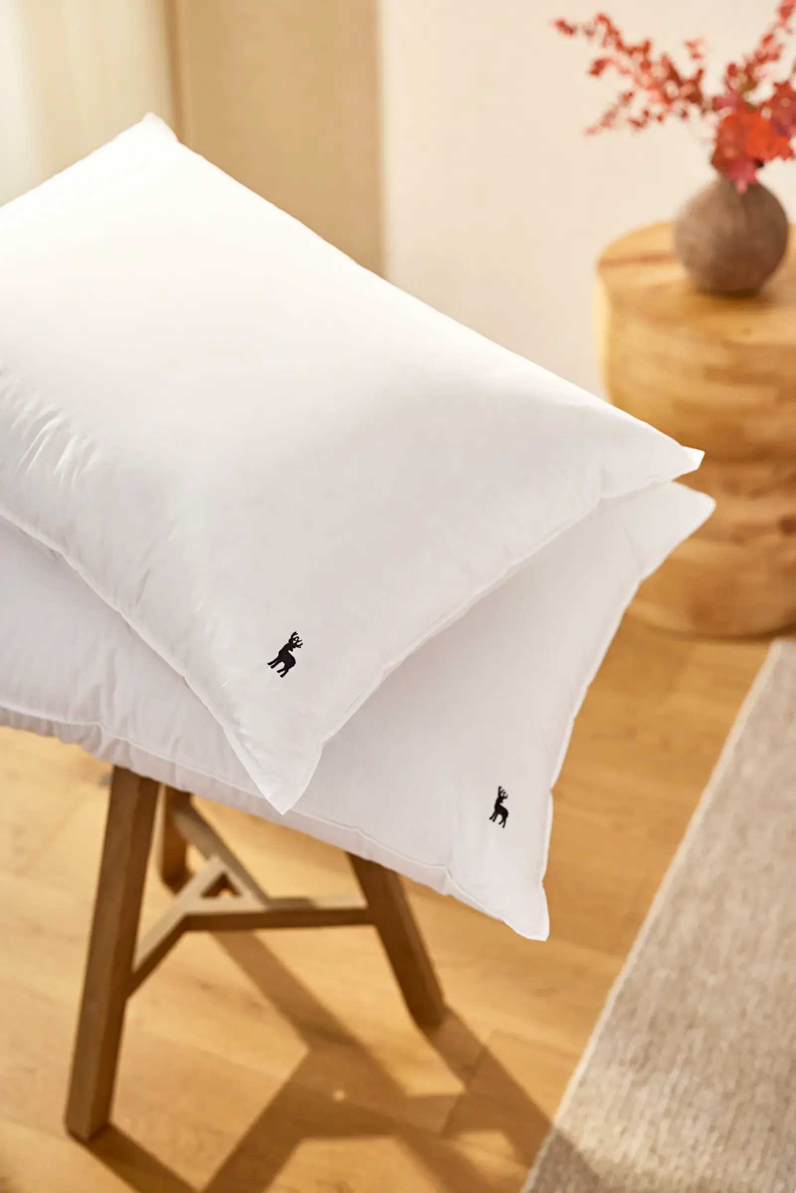 Two white pillows with small black reindeer silhouettes placed on a wooden stool in a cozy room with a wooden side table and houseplant with red leaves in a round vase.