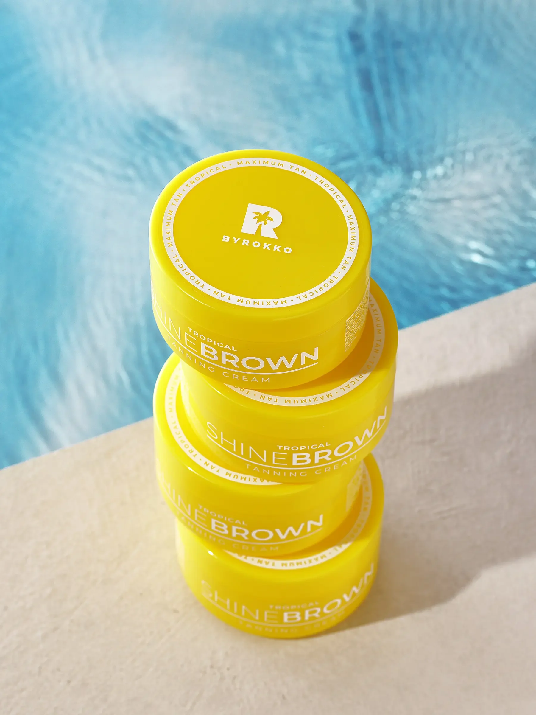 Four yellow containers of tropical tanning cream stacked vertically on a beige surface, with a pool in the background.