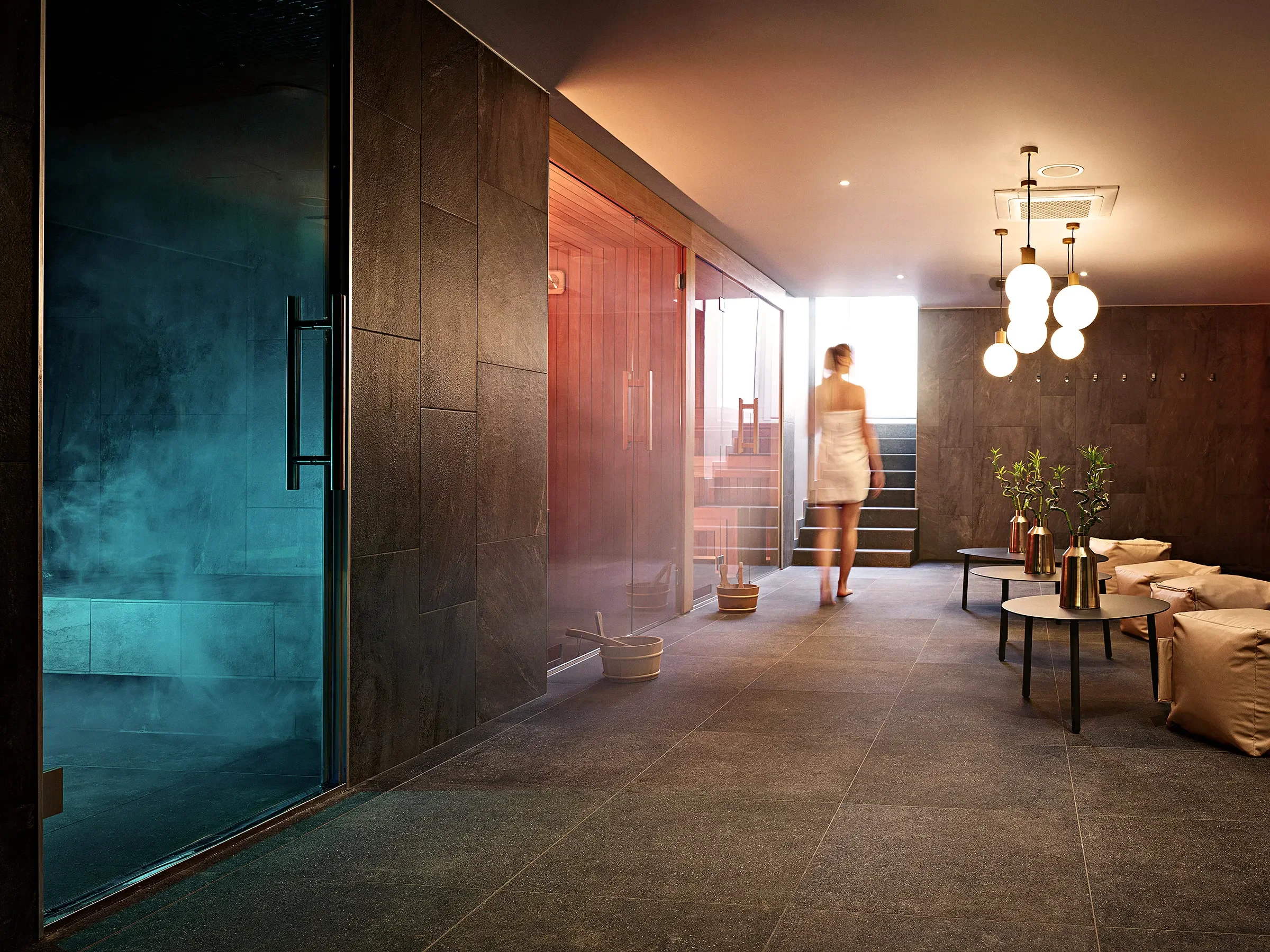 Spa lobby with steam room, seating, potted plants, and a woman in a white towel walking towards the stairs.