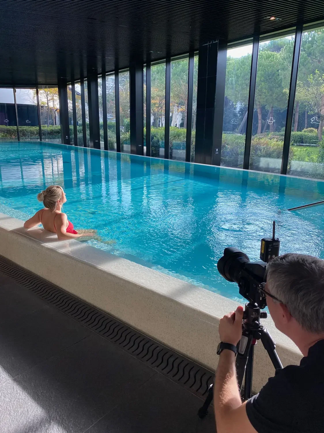 A woman in a red swimsuit sitting by the edge of an indoor swimming pool with large windows, while a photographer takes a picture of her.
