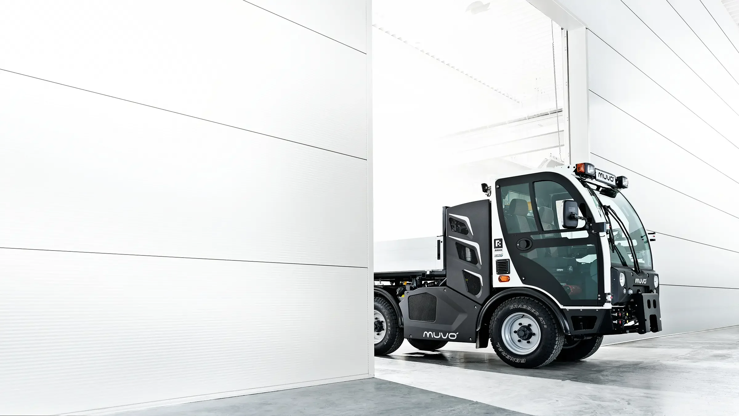 A small, modern utility vehicle with a white and black exterior parked in an industrial setting near a white wall.