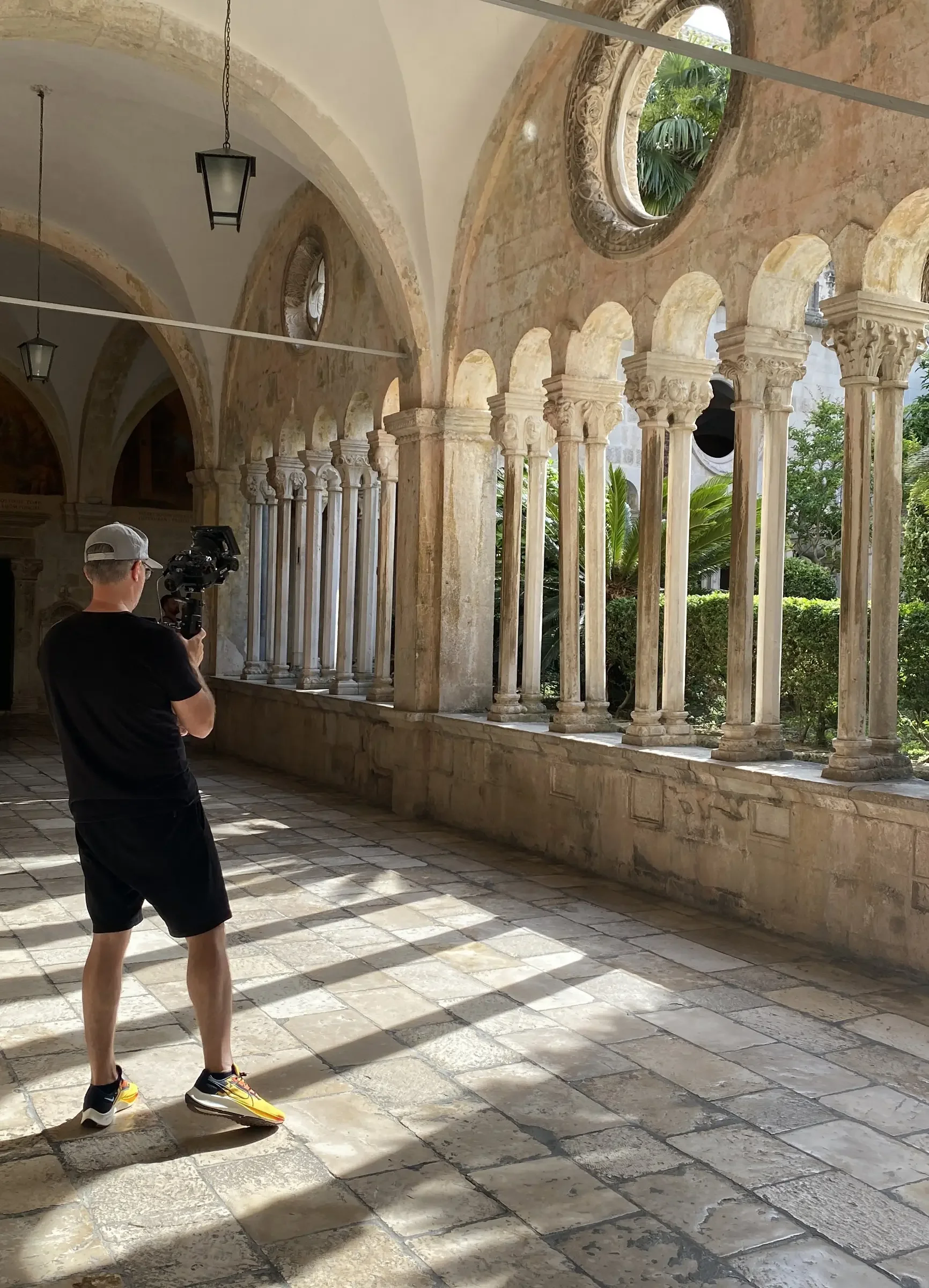 A man wearing a gray cap, black t-shirt, black shorts, and yellow sneakers is filming or taking photos with a camera on a tripod inside a historic stone building with arched windows and columns, overlooking a lush garden.