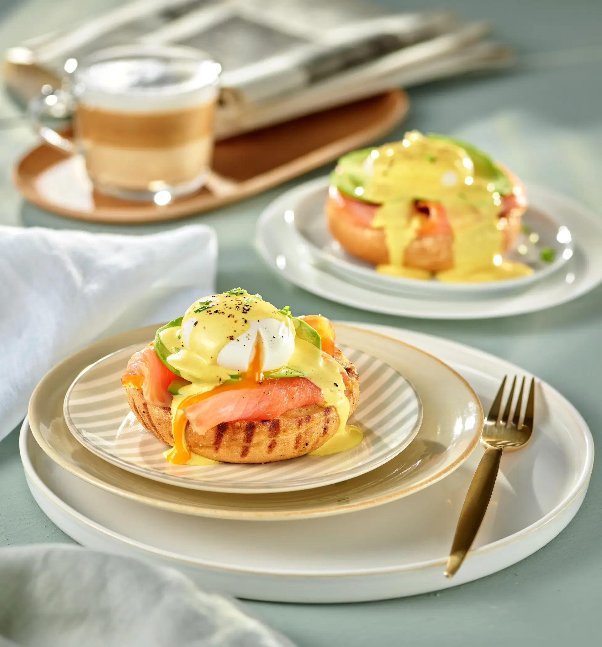 Eggs Benedict with smoked salmon, avocado slices, and hollandaise sauce served on a grilled English muffin with a poached egg on top, plated on a beige and white dish, with a fork on a white tray, and another similar dish in the background.