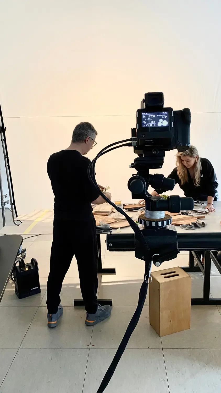 Two people working at a table with food, a camera on a tripod is recording them, in a studio setting.