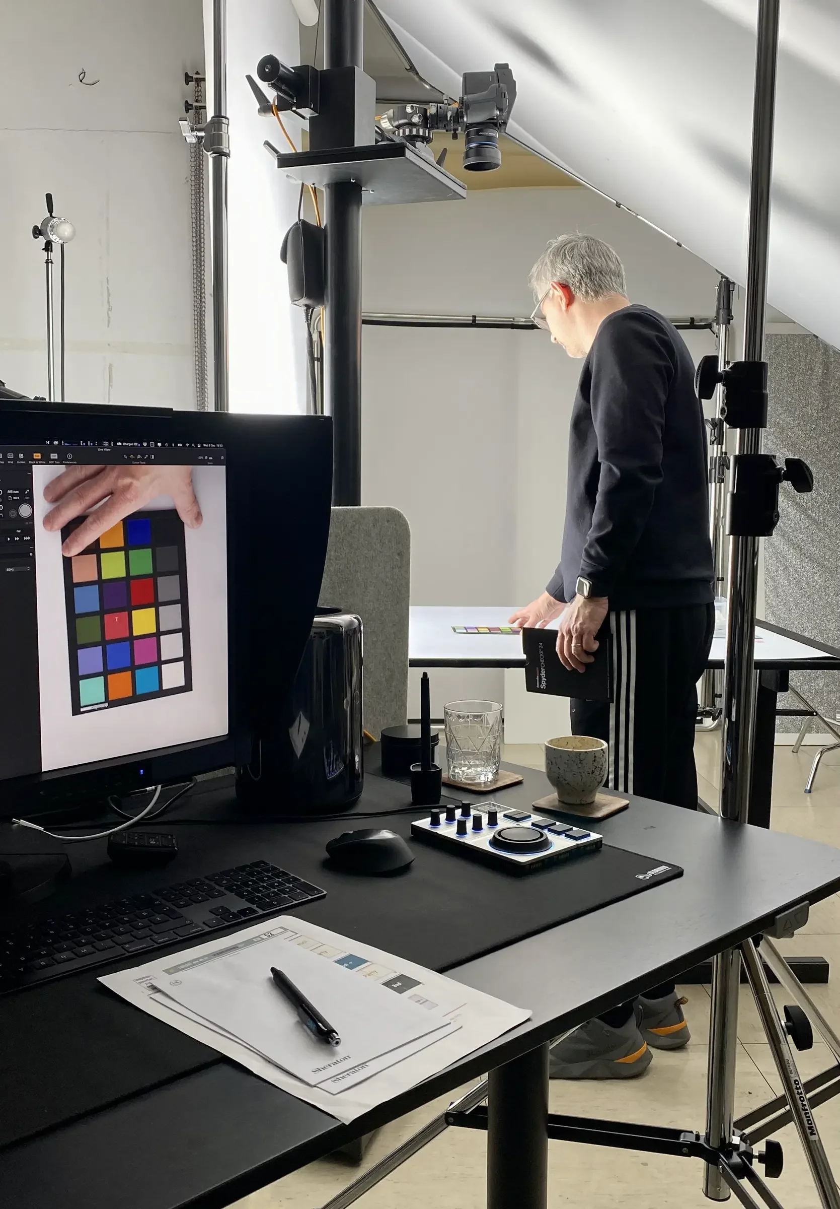 A man wearing black sweater and striped pants standing in a photography studio, looking at a color chart on a table, surrounded by photography equipment and a computer monitor displaying a color test chart.