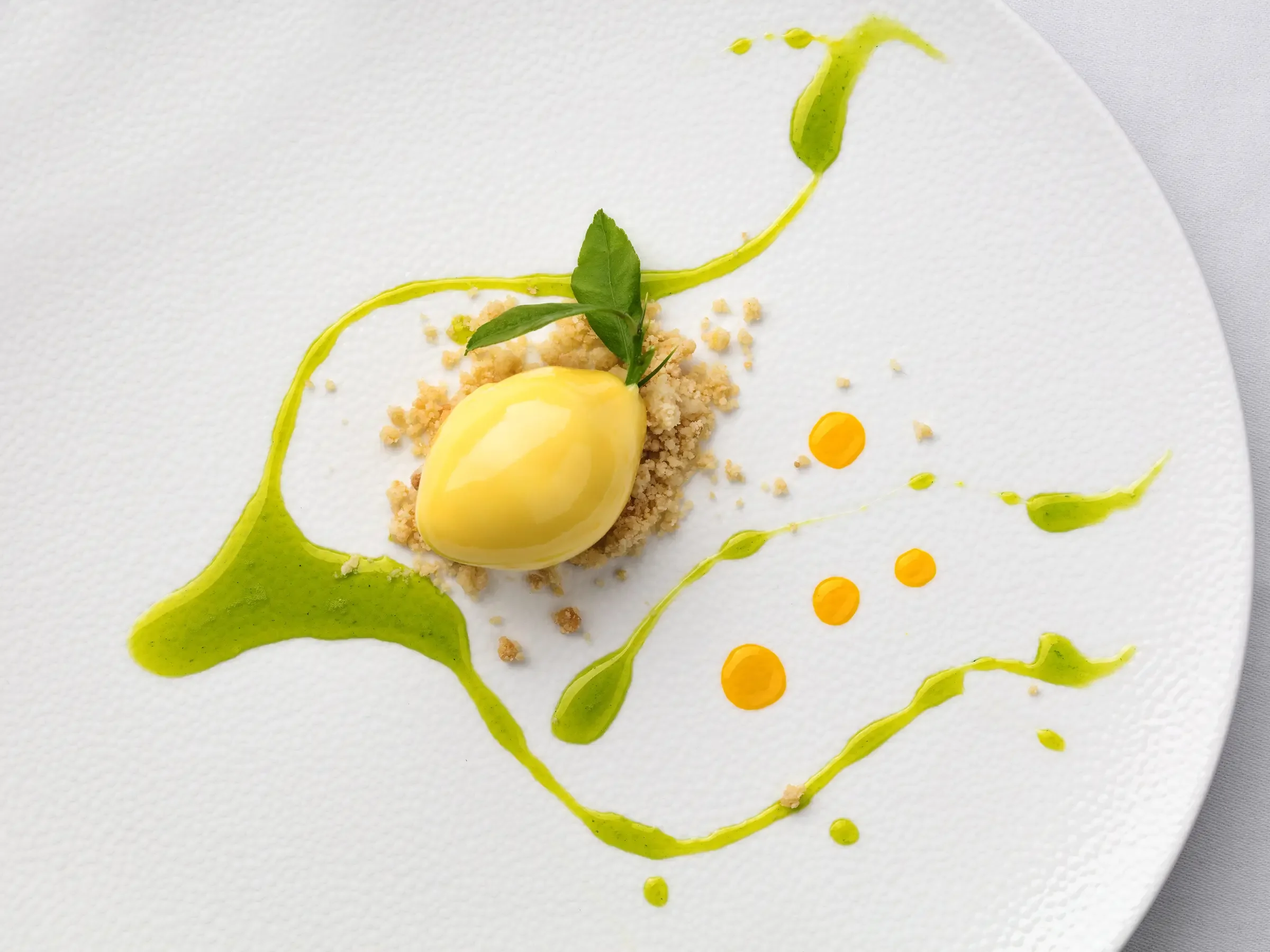 A gourmet dessert featuring a yellow quenelle of mousse or sorbet, garnished with green leaves, on a bed of crumbled biscuit or cake crumbs, with green and orange sauce drizzled artistically on a white textured plate.