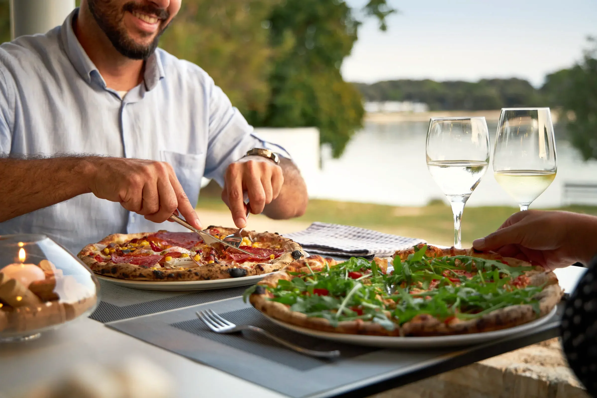 People enjoying pizza outdoors with a lake in the background, two glasses of white wine on the table.