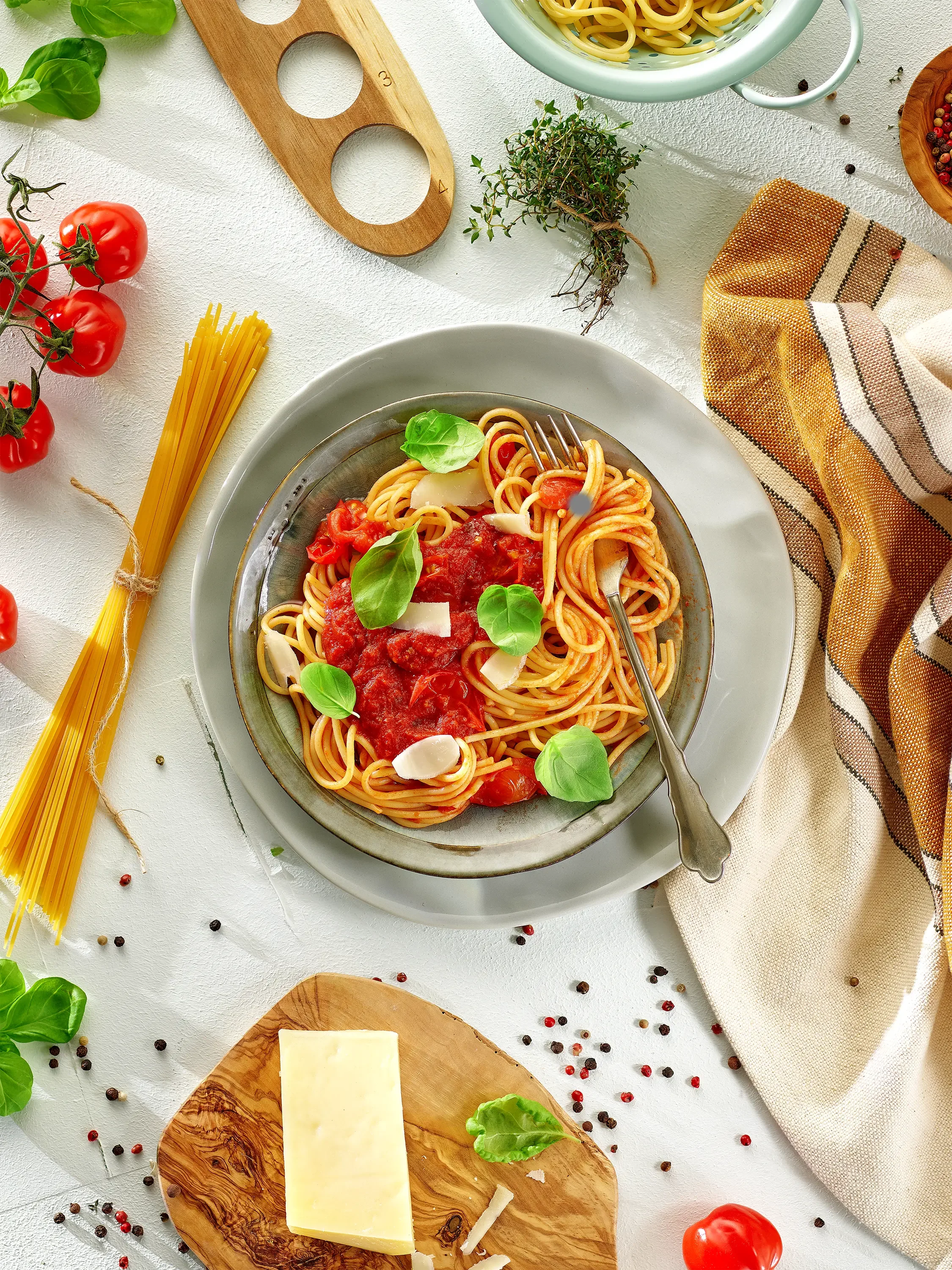 A plate of spaghetti with tomato sauce garnished with basil leaves and Parmesan cheese, surrounded by cherry tomatoes, dried pasta, herbs, peppercorns, and a block of cheese on a wooden cutting board.